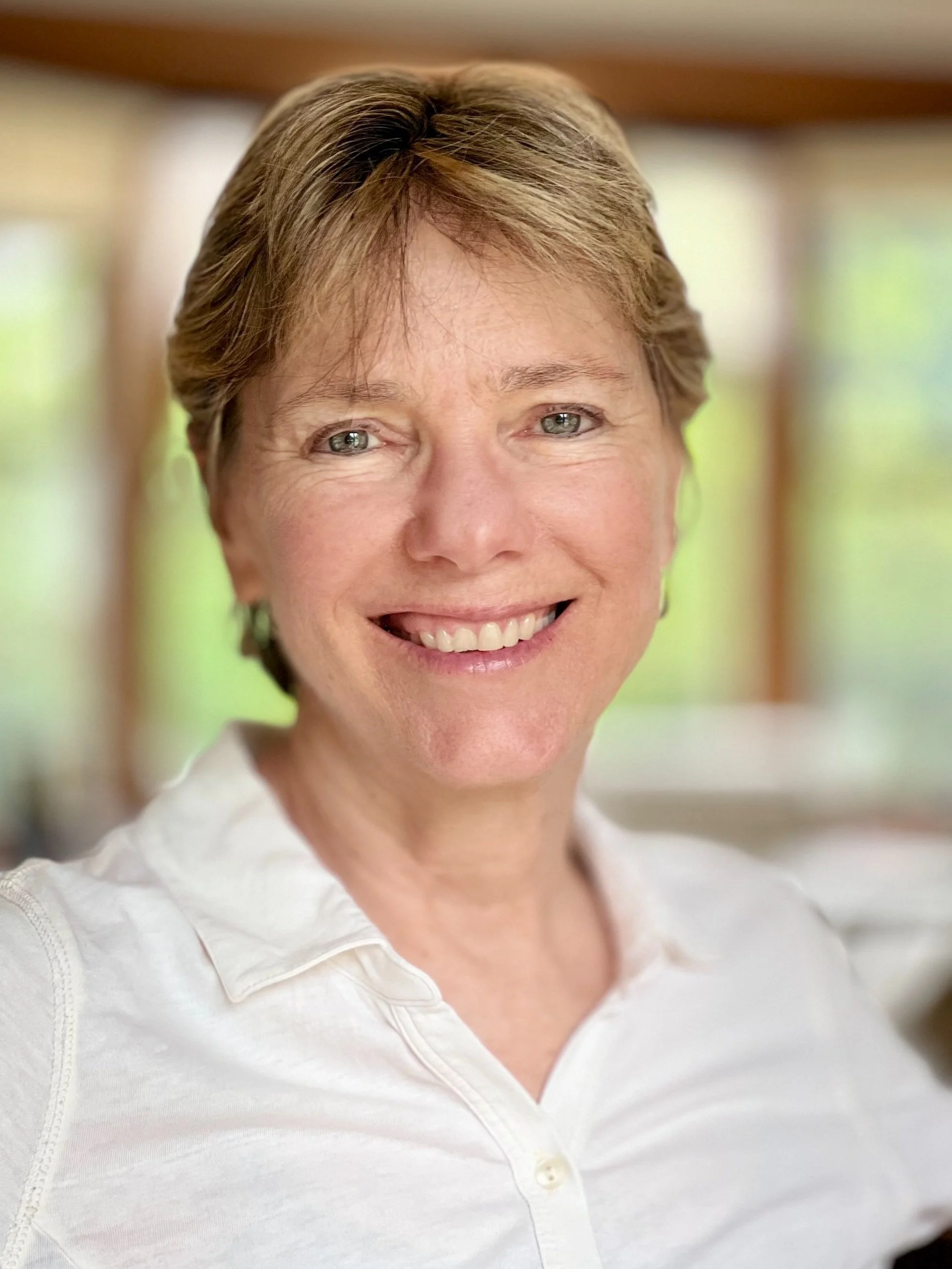 Close-up of a smiling woman with short brown hair, wearing a white collared shirt, standing indoors with a blurred background of windows and natural light.