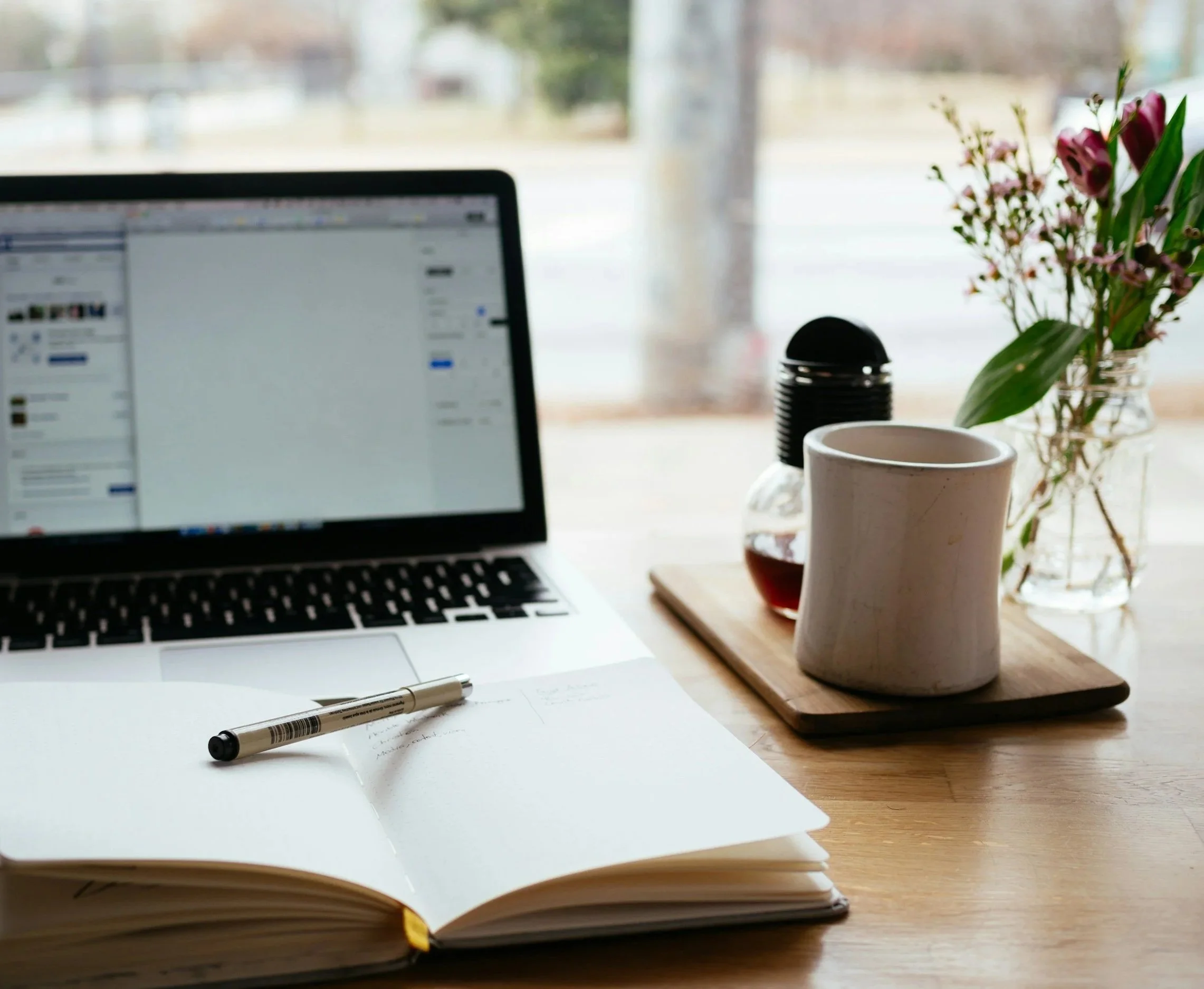 Desk in a therapist office, with an open notebook of handwritten notes, a black marker, a pile of papers, a pen, a computer, a cup on a wooden tray, and a vase with pink flowers on a wooden table near a window.