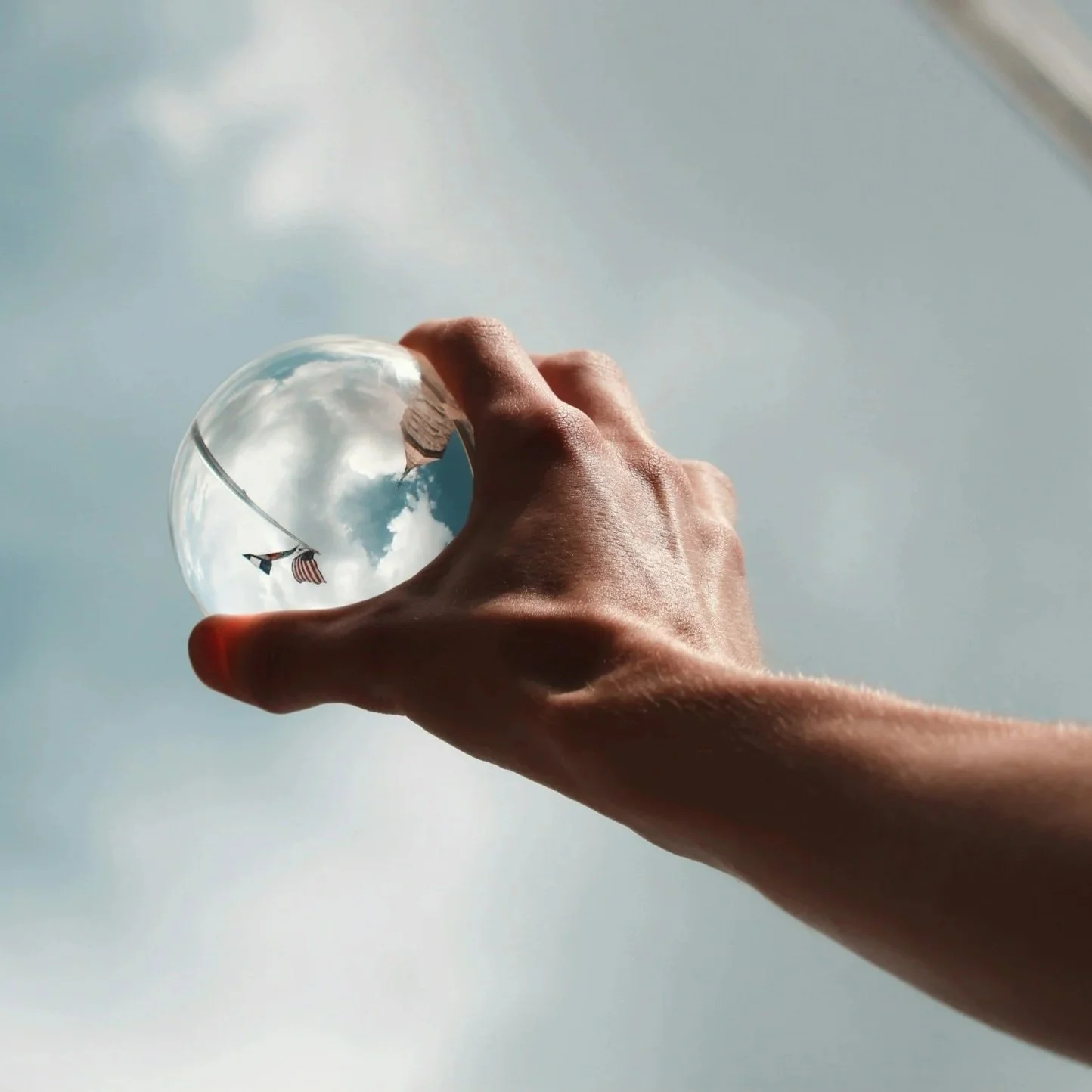 A hand holding a small glass ball, reflecting a cloudy sky and flags in the background.