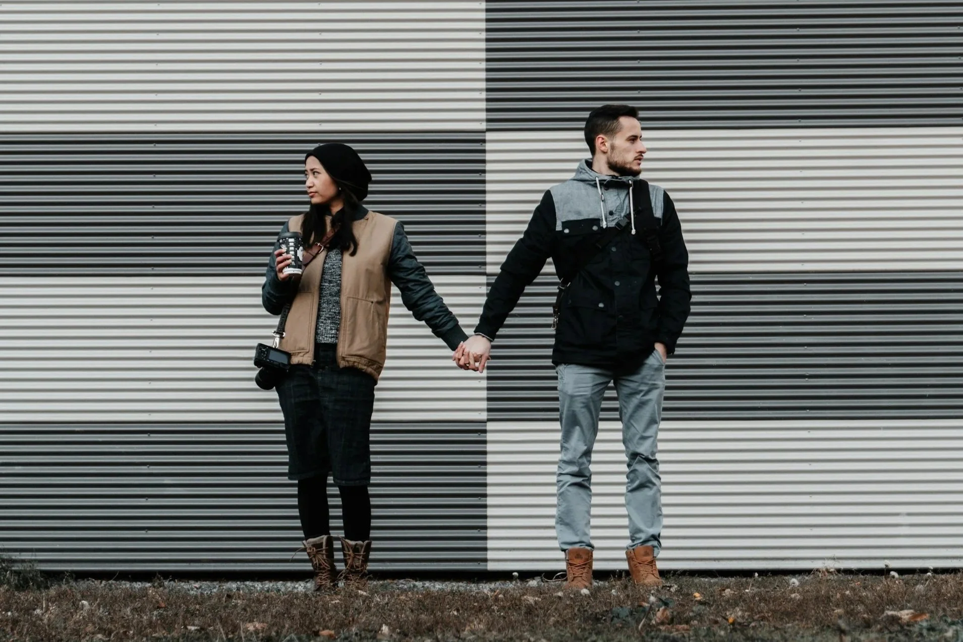 A young man and woman holding hands, standing against a large corrugated metal wall with horizontal stripes. The woman has long dark hair, a beige vest, gray sweater, checkered pants, and brown boots, holding a coffee cup and a camera. The man has short dark hair, a black and gray jacket, gray pants, and tan boots, with one hand in his pocket and the other holding the woman's hand.