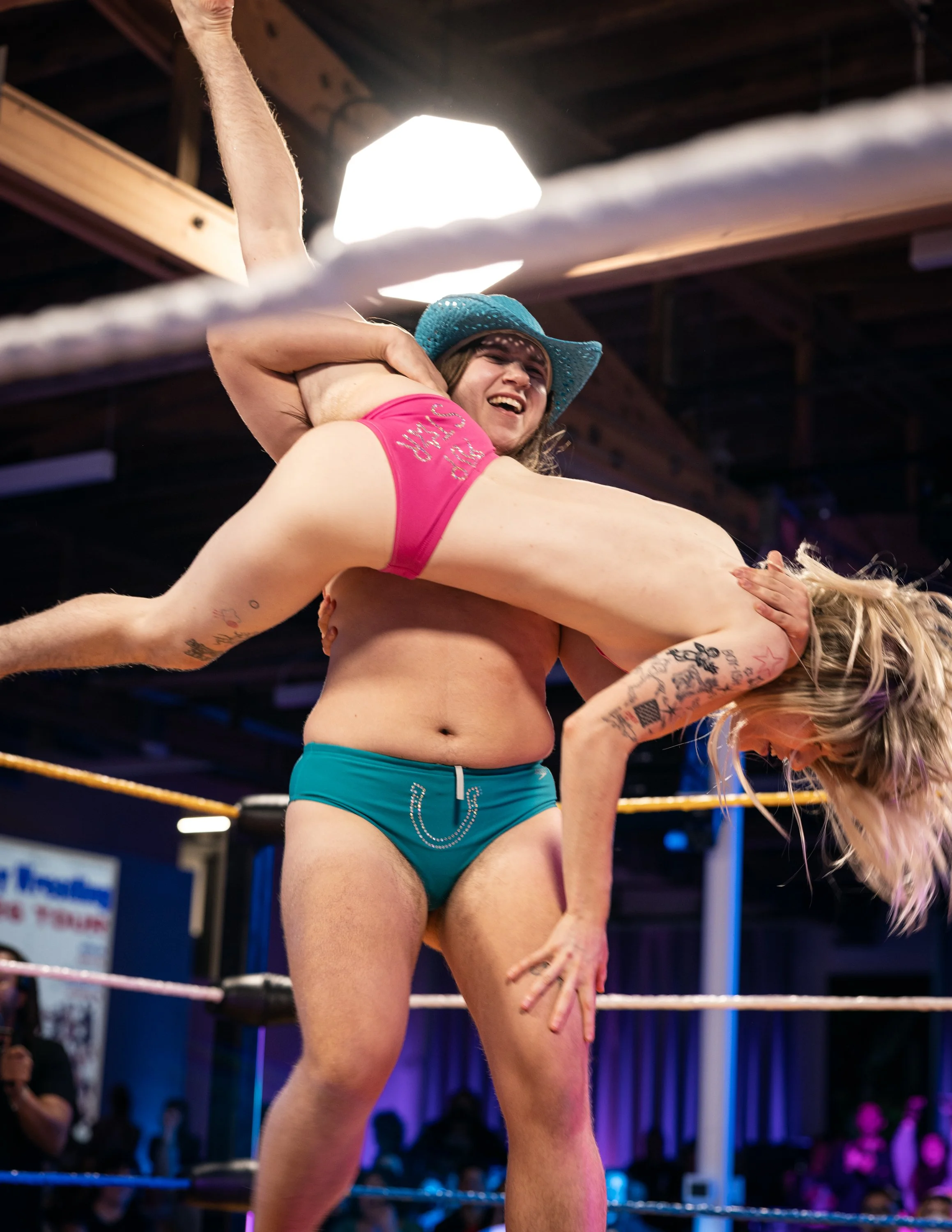 A trans wrestler lifts up their opponent in preparation for a body slam in PDX for TBOY Wrestling.