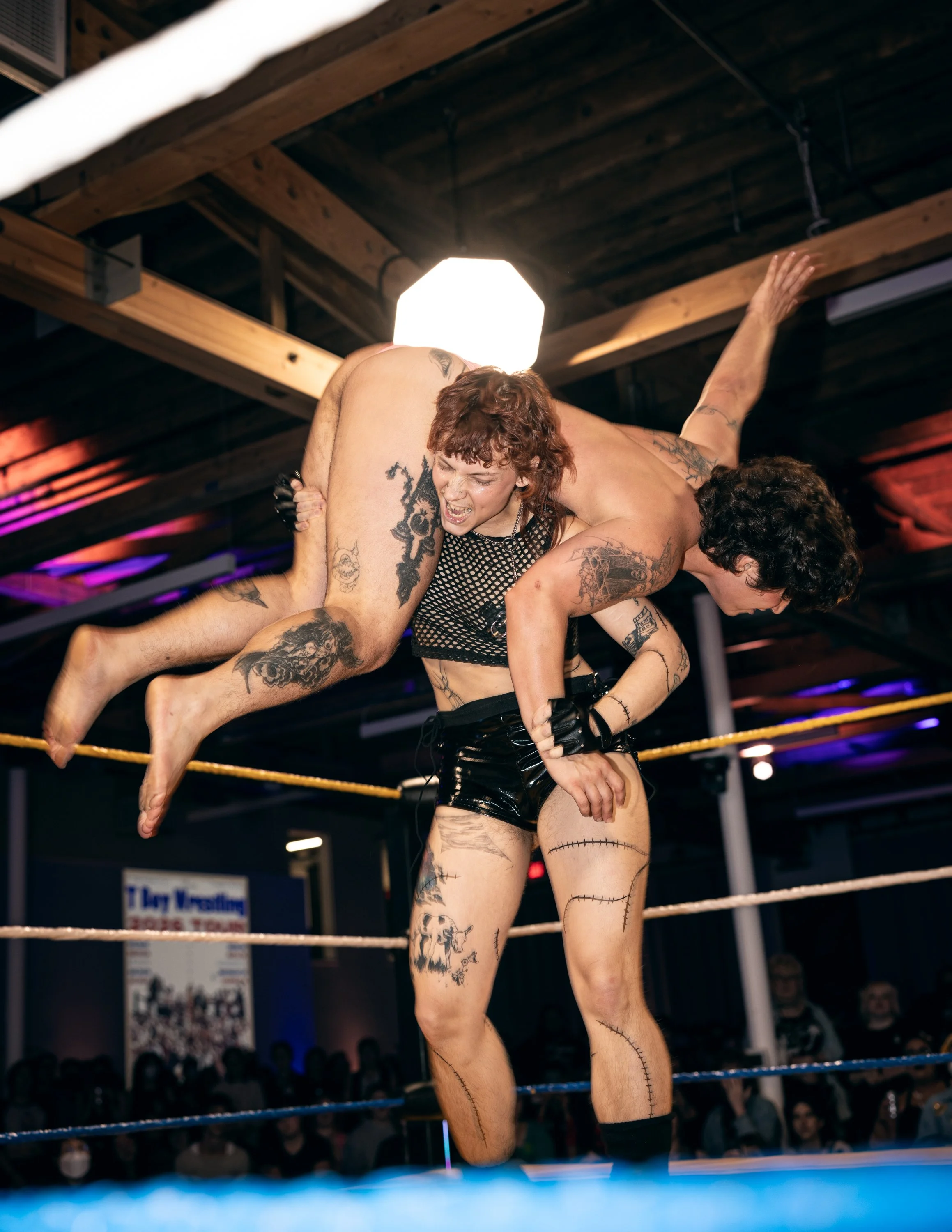 A trans wrestler lifting their opponent on their back in preparation for a brutal body slam at TBOY Wrestling in Portland, OR.