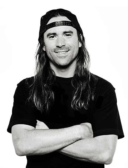 Portrait of a man with long hair, wearing a backward baseball cap and a black short-sleeve shirt, smiling with arms crossed.