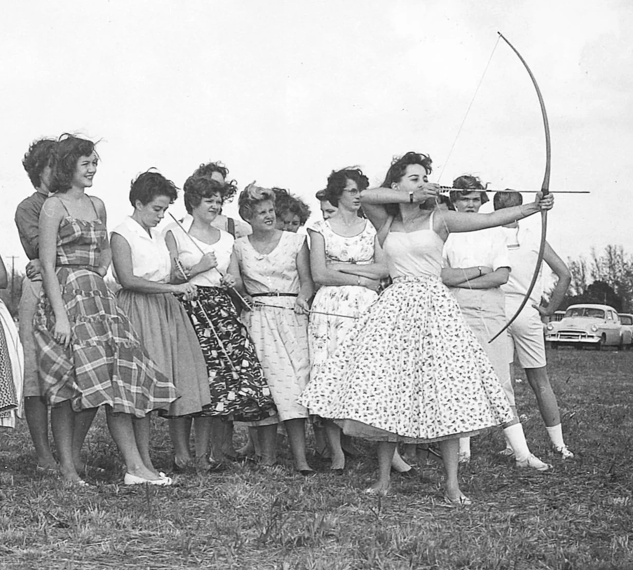 Group of women in mid‑century dresses gathered on a grassy field as one draws a bow, capturing teamwork, focus, and vintage field‑day energy.