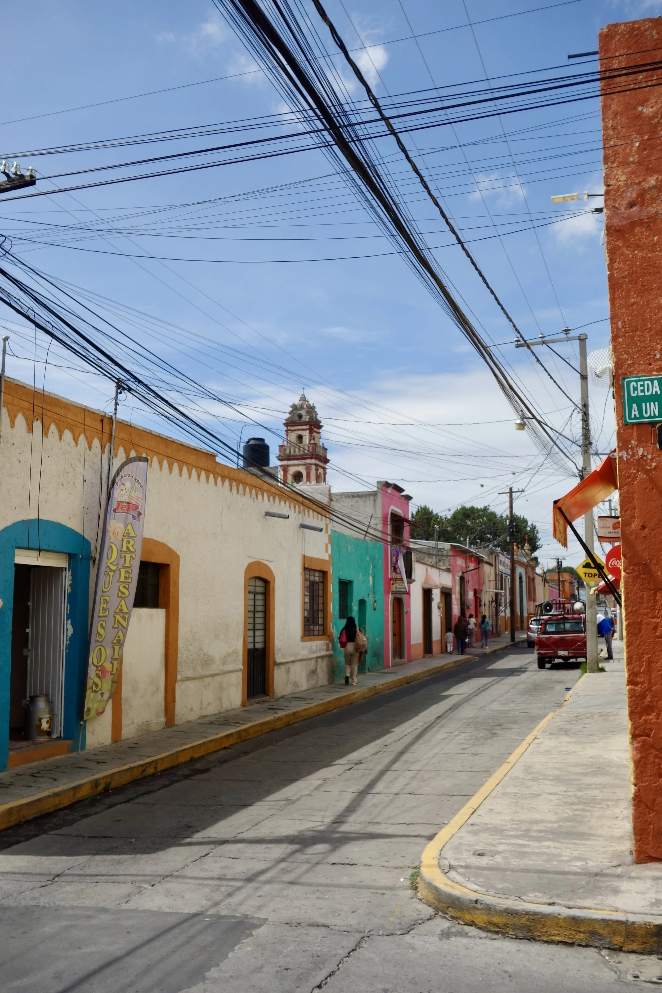 Colorful street in Huamantla, Tlaxcala, Mexico, with houses painted in various bright colors, overhead electrical wires, pedestrians walking, and a church tower.