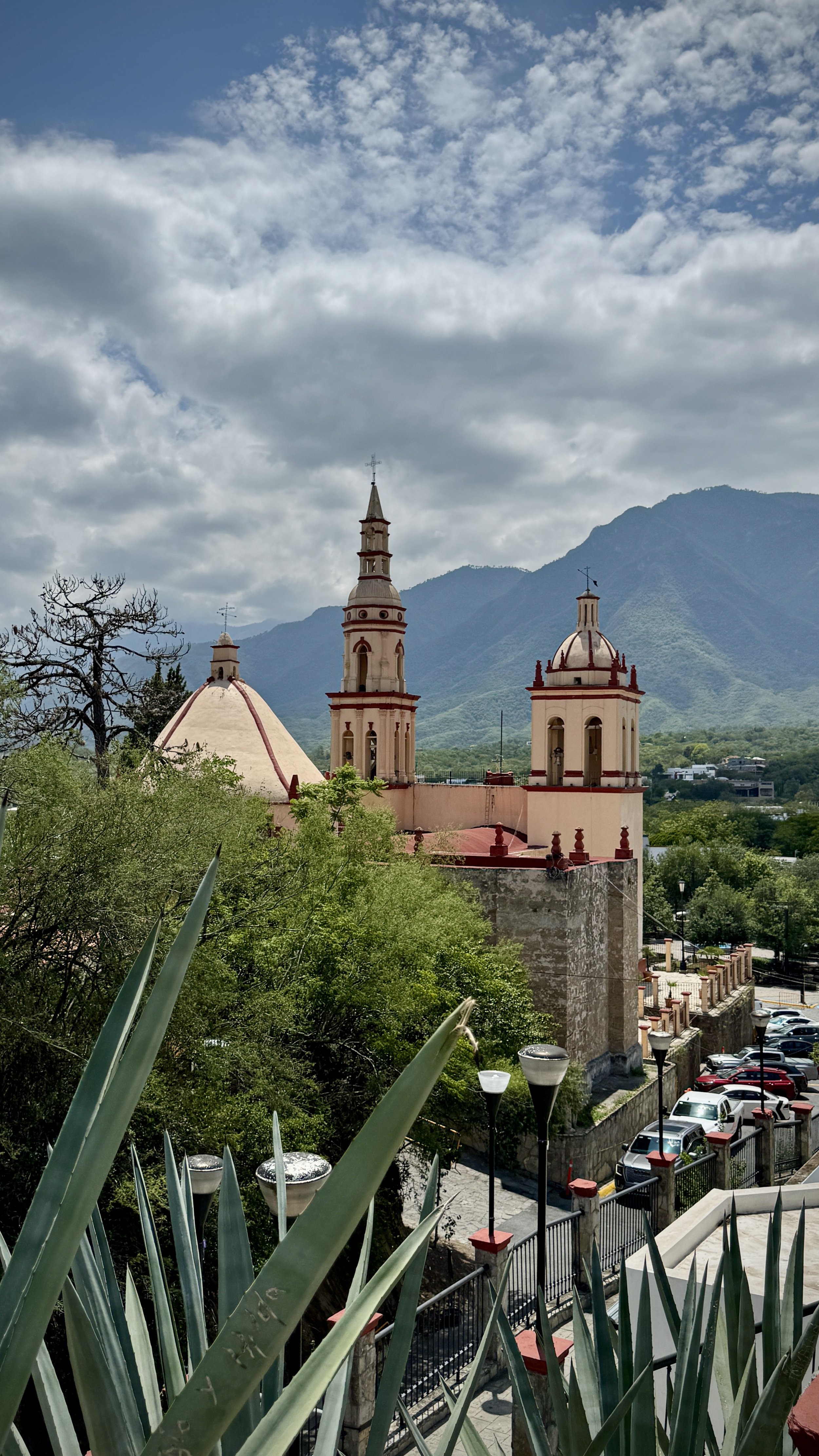 A church with pink walls and three steeples surrounded by greenery, with mountains in the background and a partly cloudy sky overhead in Santiago, Nuevo Leon, Mexico.