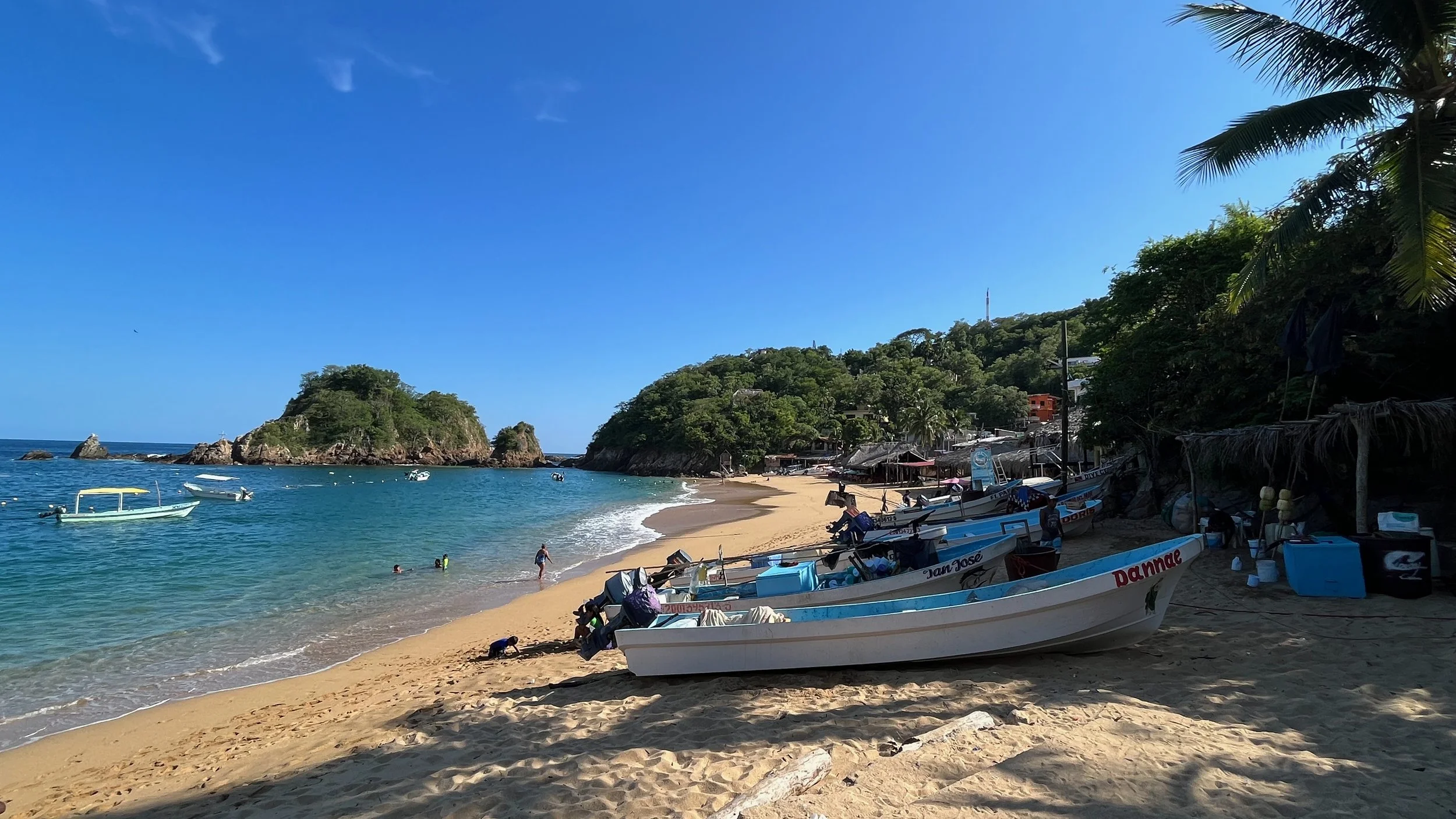 A sandy beach with several small boats anchored along the shore, clear blue water, a few people swimming, and green hills with trees in the background under a bright blue sky in Puerto Angel, Oaxaca, Mexico.