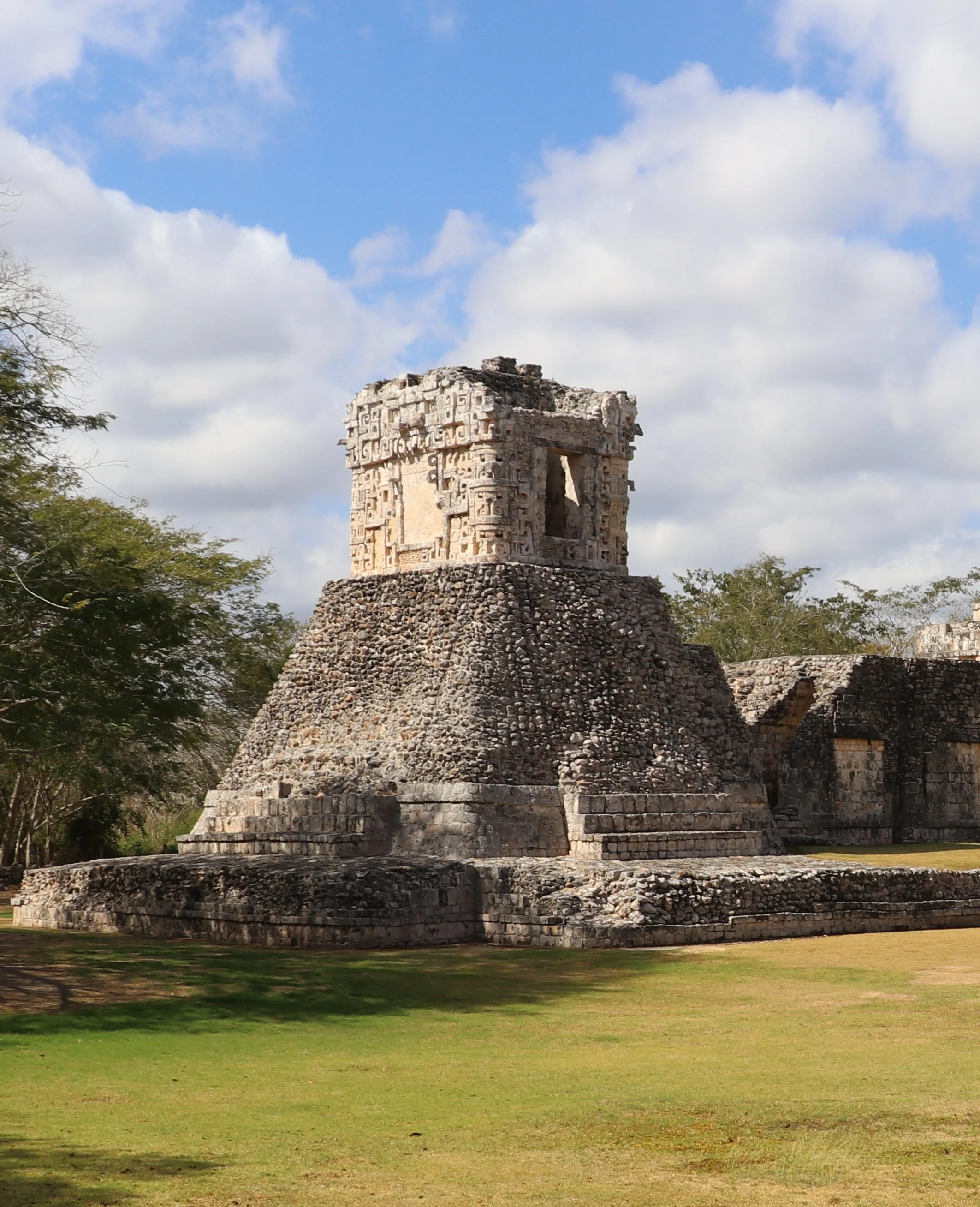 A partial view of the ancient Maya temple of Dzibilnocac in the Chenes region of Campeche, Mexico, set in a grassy area under a partly cloudy sky.
