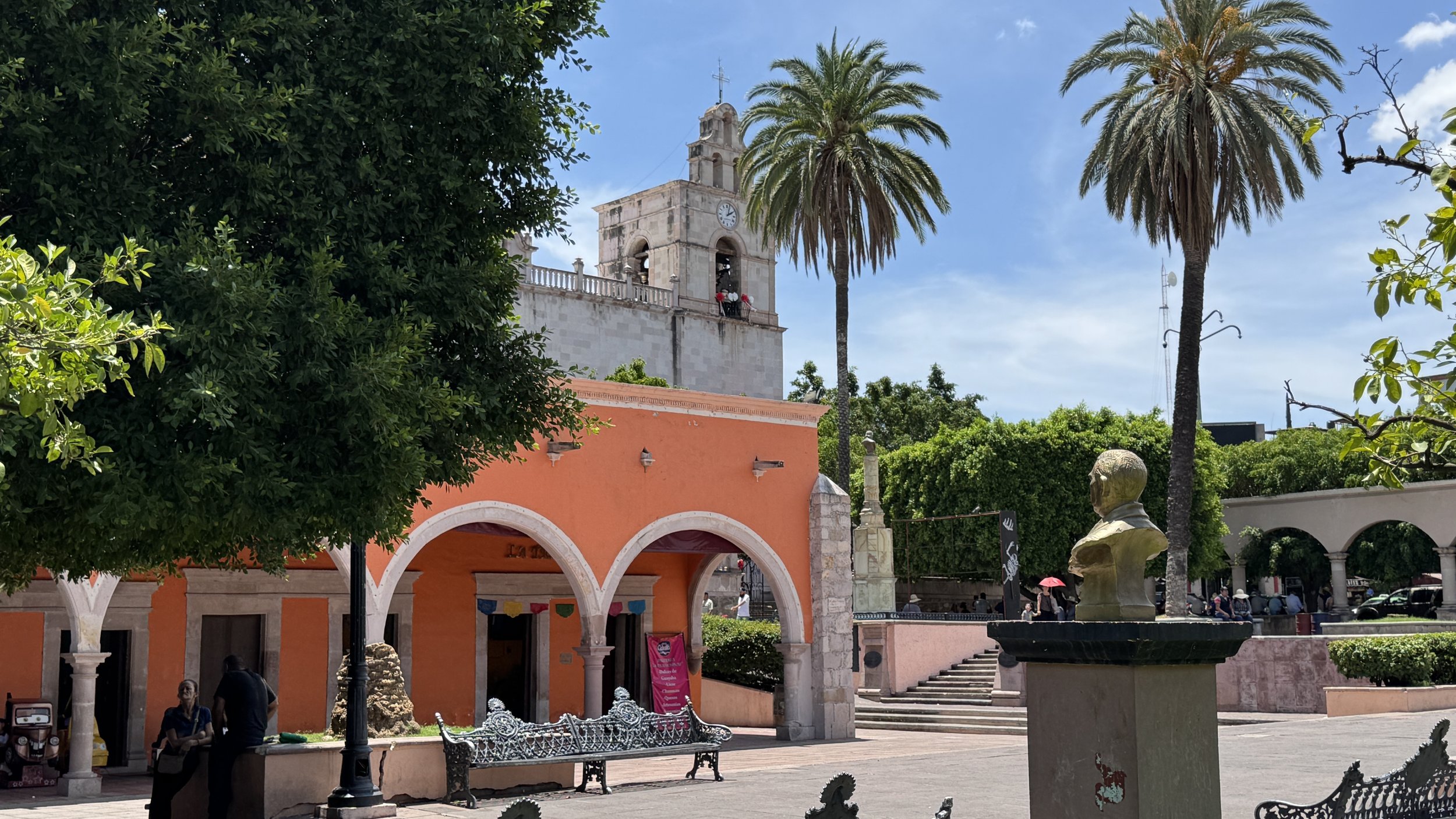 The zocalo, with an orange building with archways, green trees, tall palm trees, statues, benches, people, and a clock tower in the background under a blue sky in Calvillo, Aguascalientes, Mexico.