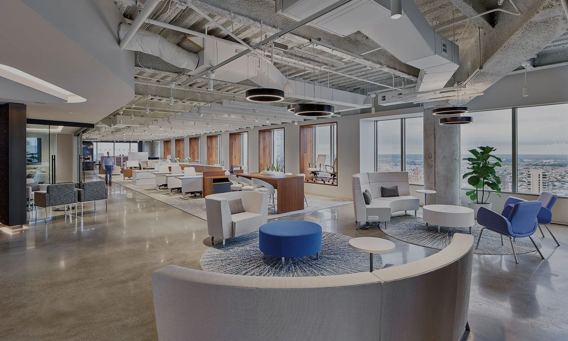 Modern open office space with large windows, lounge seating, desks, and overhead lighting.