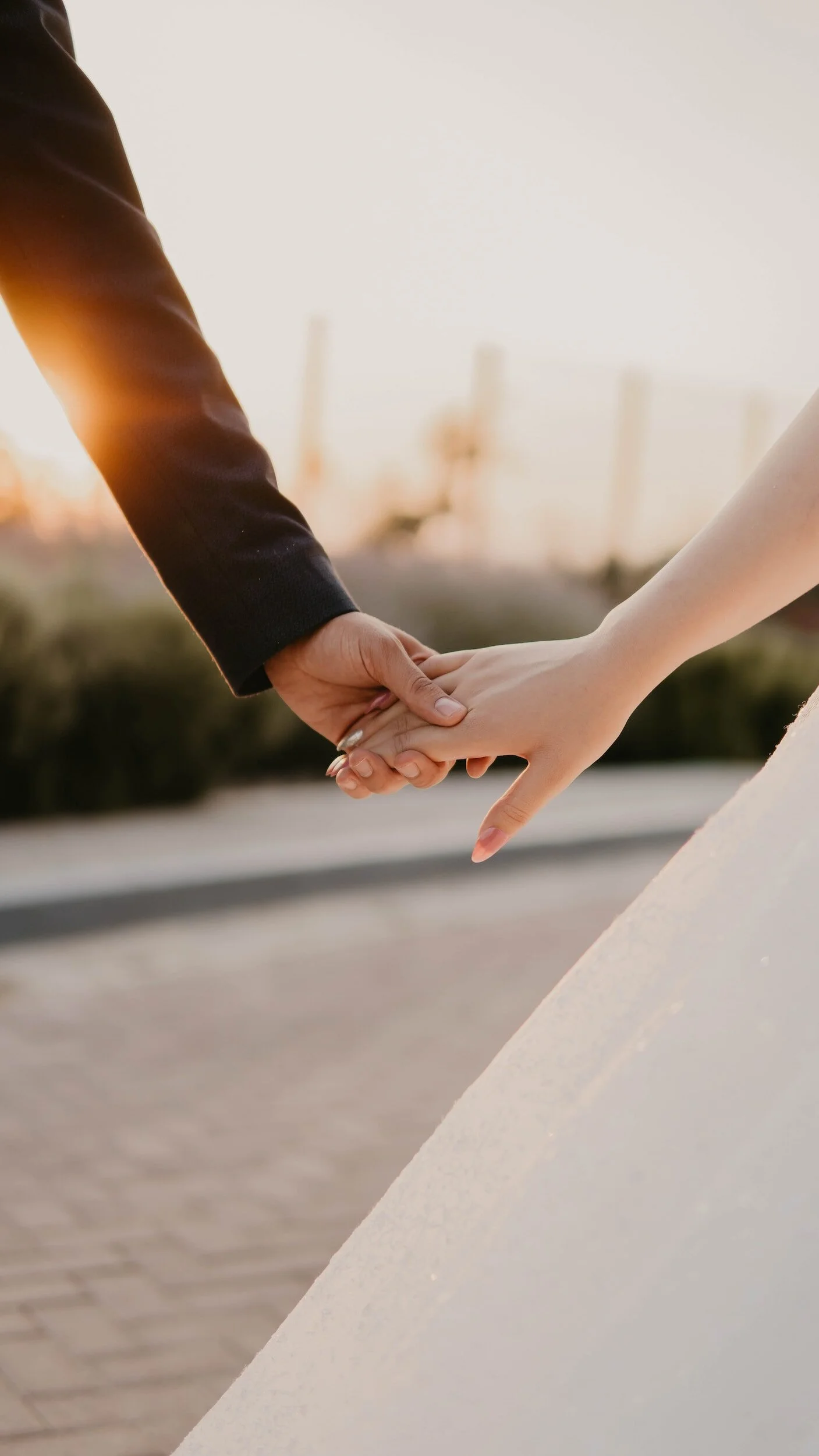 A man and woman holding hands, with the woman's hand resting on top of the man's hand, outdoors during sunset.