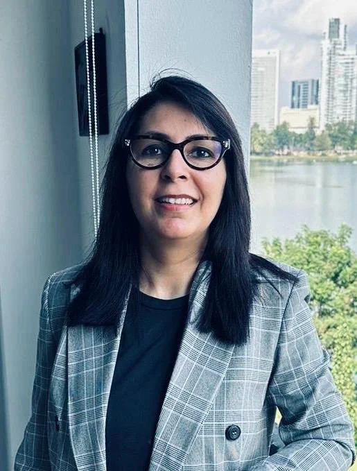 A woman with long dark hair, glasses, and fair skin wearing a white button-up shirt and dark blue skirt, standing against a dark gray background.
