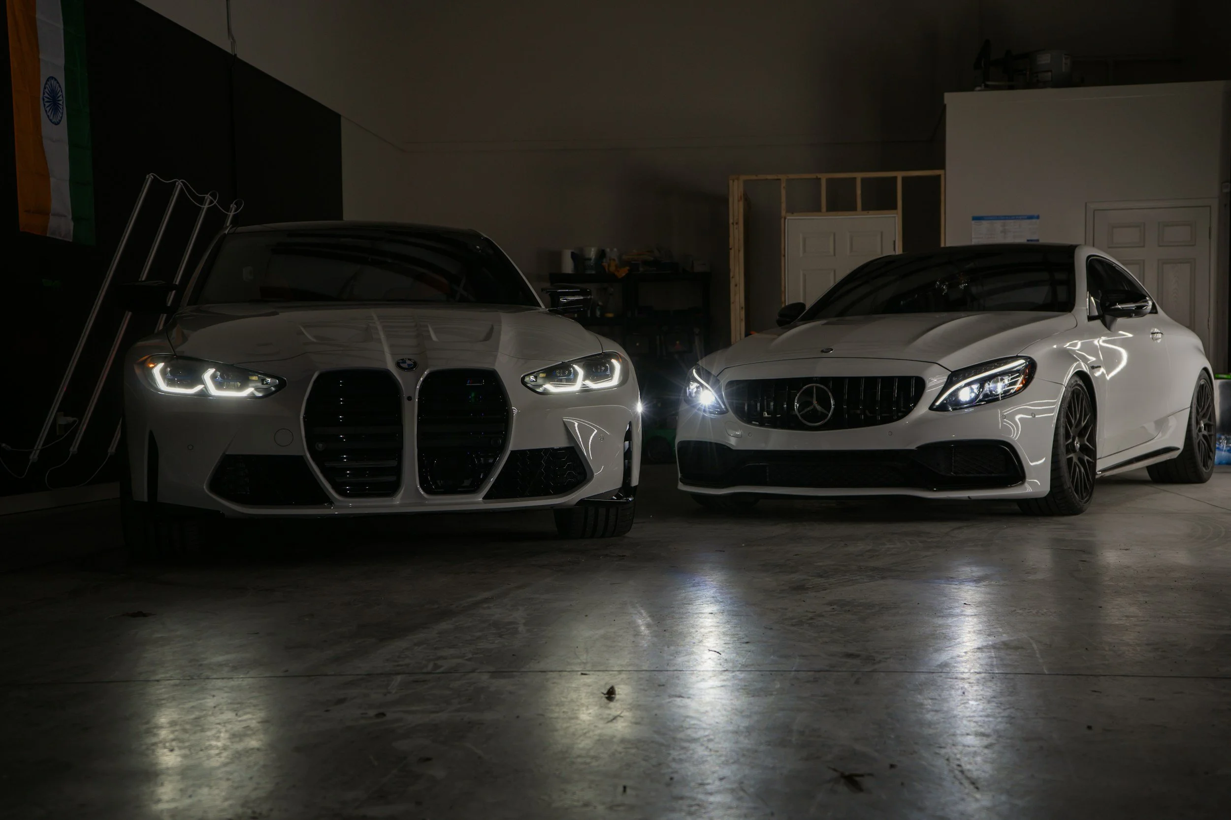 Two white luxury cars, a BMW and a Mercedes-Benz, parked in a dimly lit garage with their headlights on.