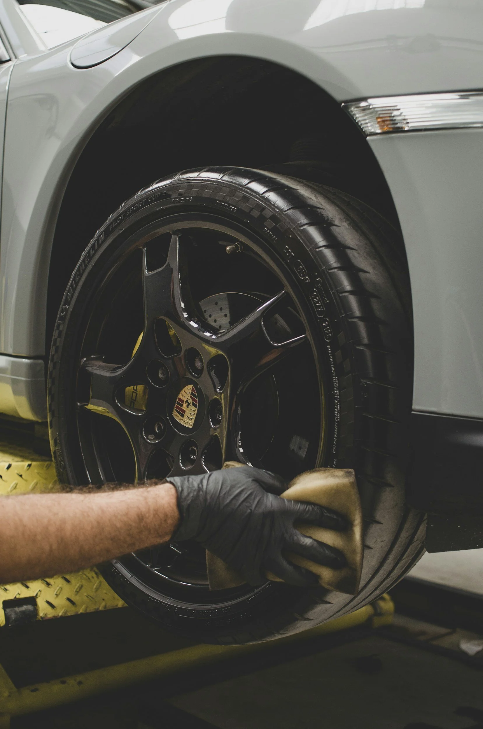 Close-up of a person wearing black gloves polishing the wheel of a silver sports car with a cloth, on a yellow automotive lift.