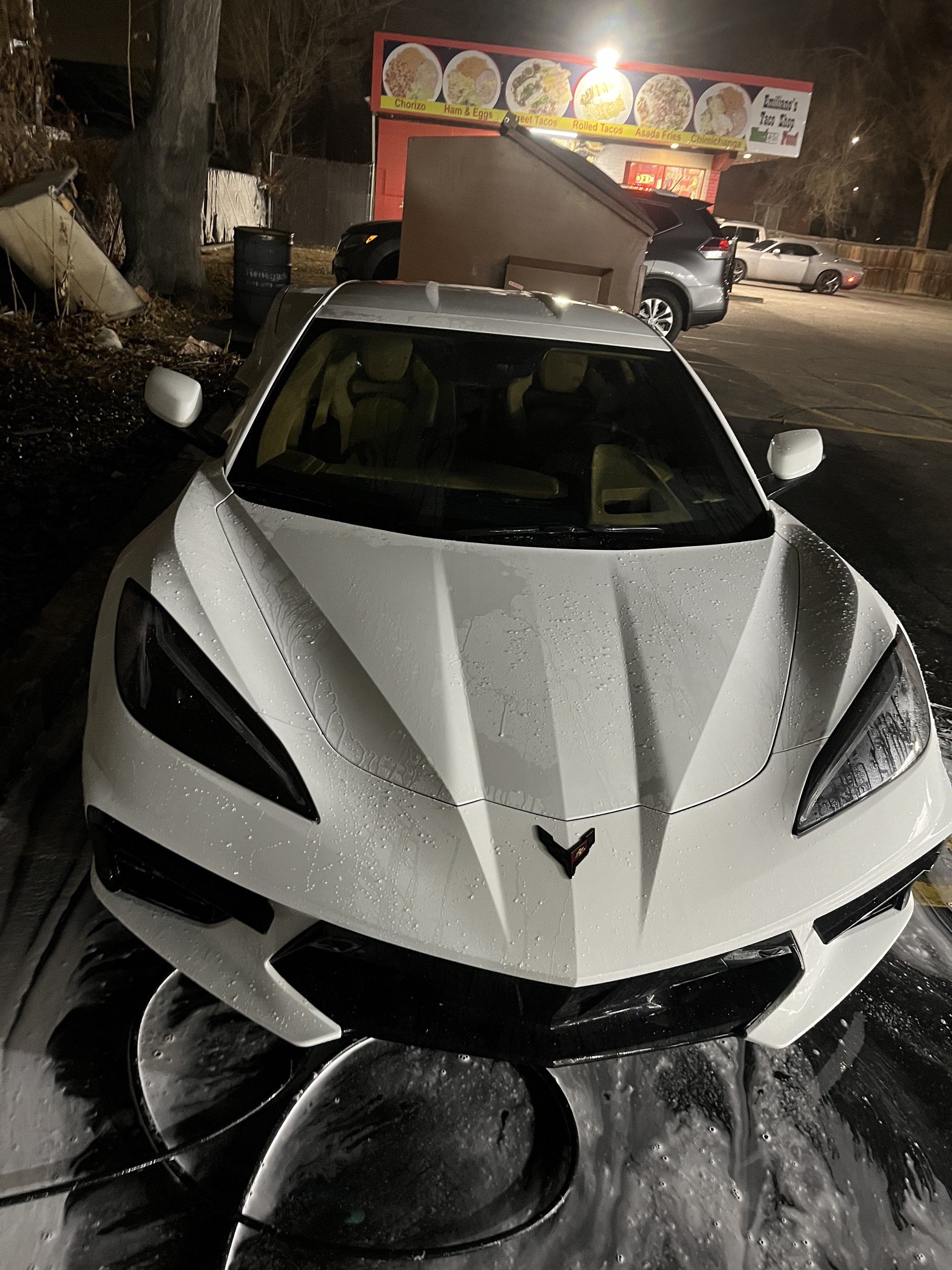 A white Corvette sports car is being washed with water and soap at night in a parking lot.