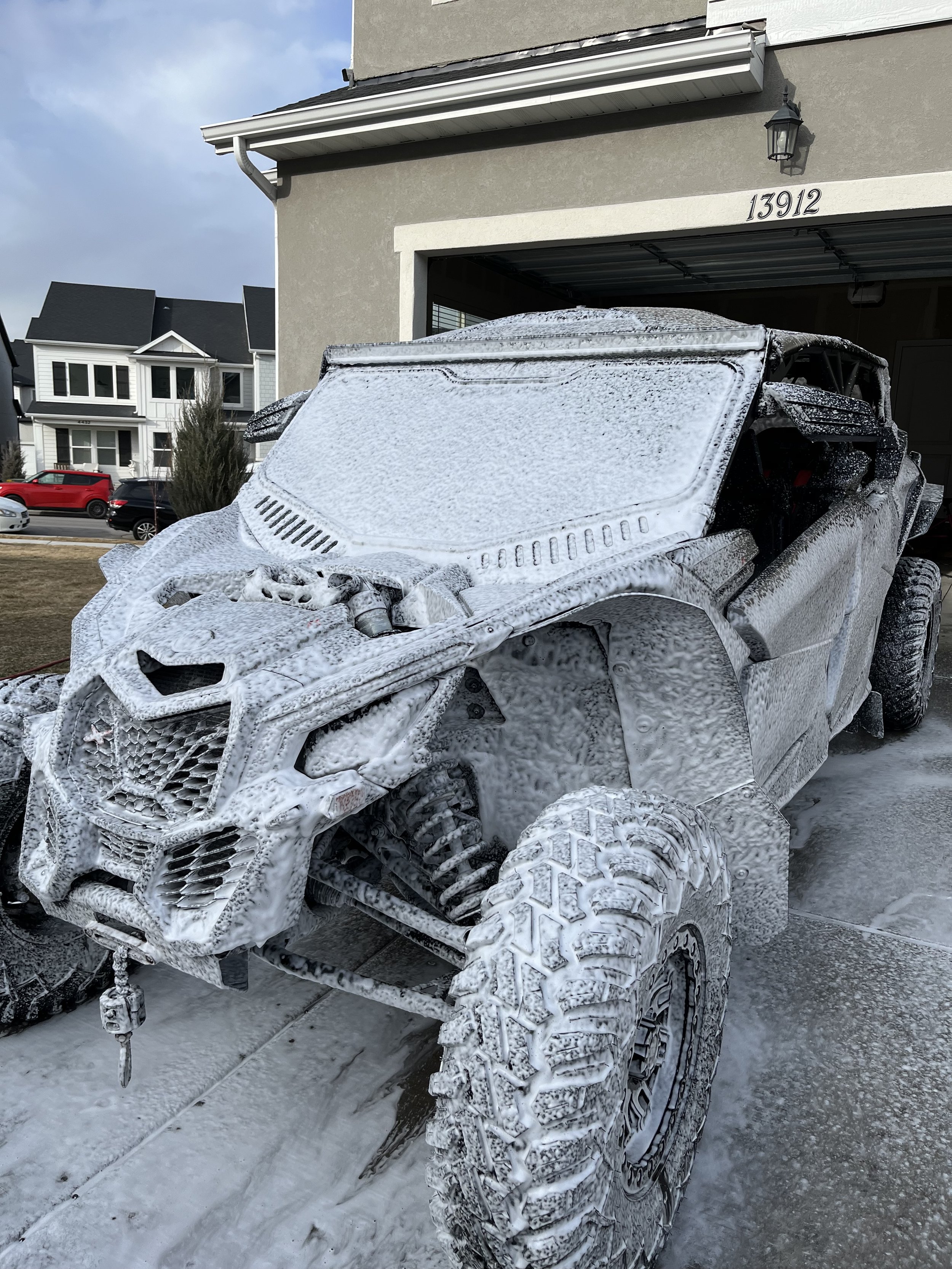 A rugged off-road vehicle covered in soap and foam in a driveway, with a house and cars in the background.
