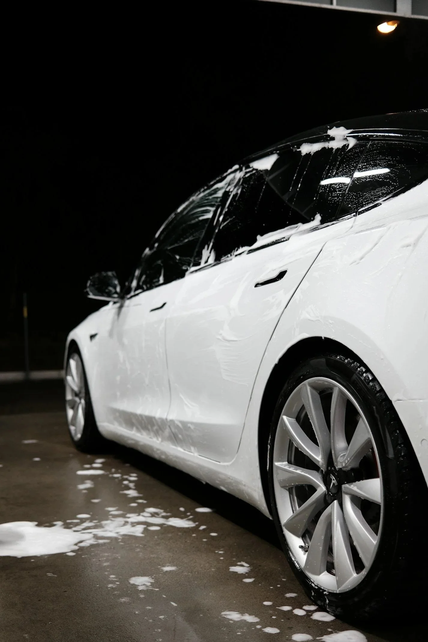 A white Tesla car at a car wash with soap suds on its body and wheel, under bright overhead lighting at night.