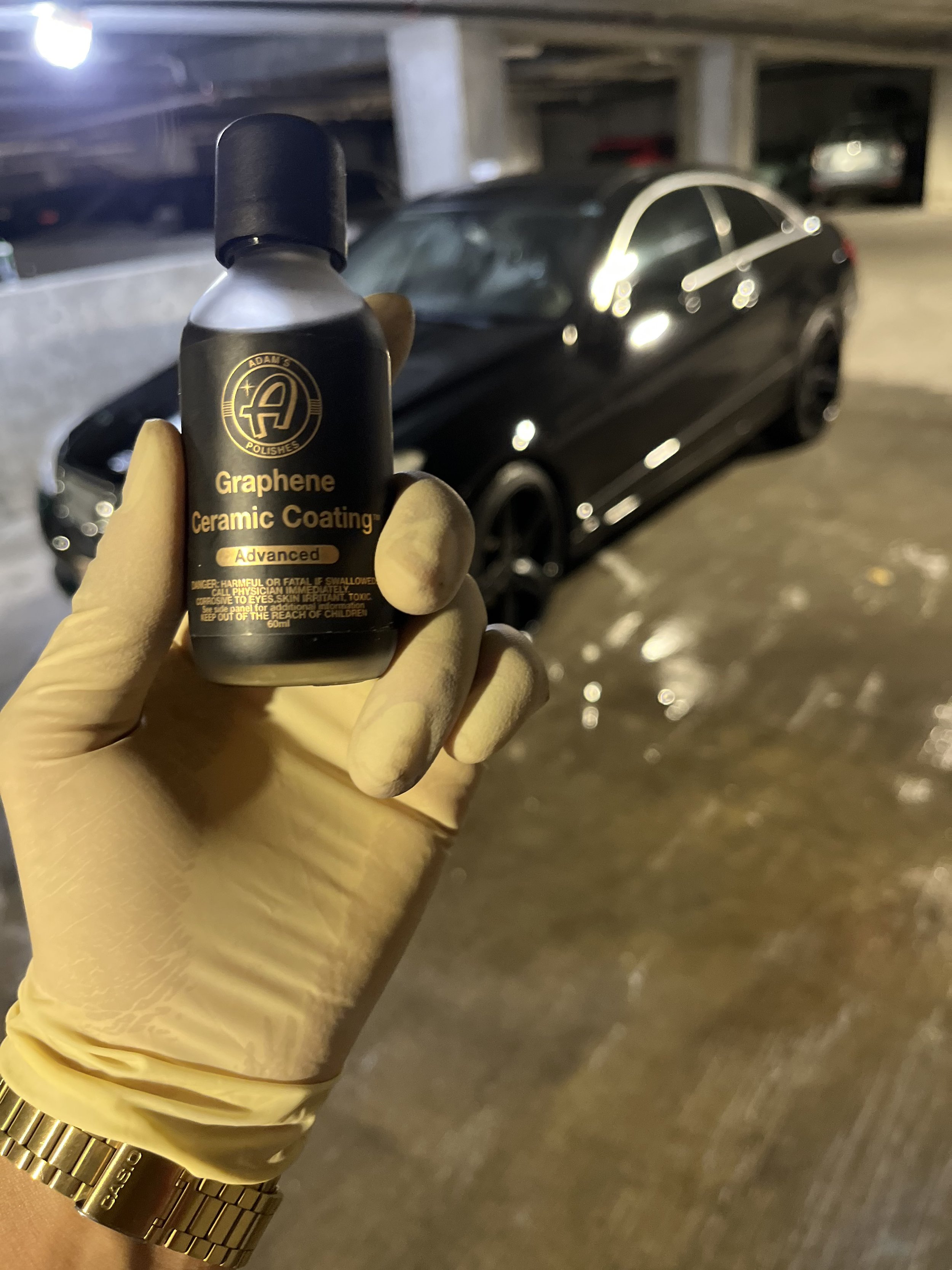 A person wearing a yellow glove and a gold watch holding a bottle of Adams Polishes Graphene Ceramic Coating in front of a black car in a parking garage.