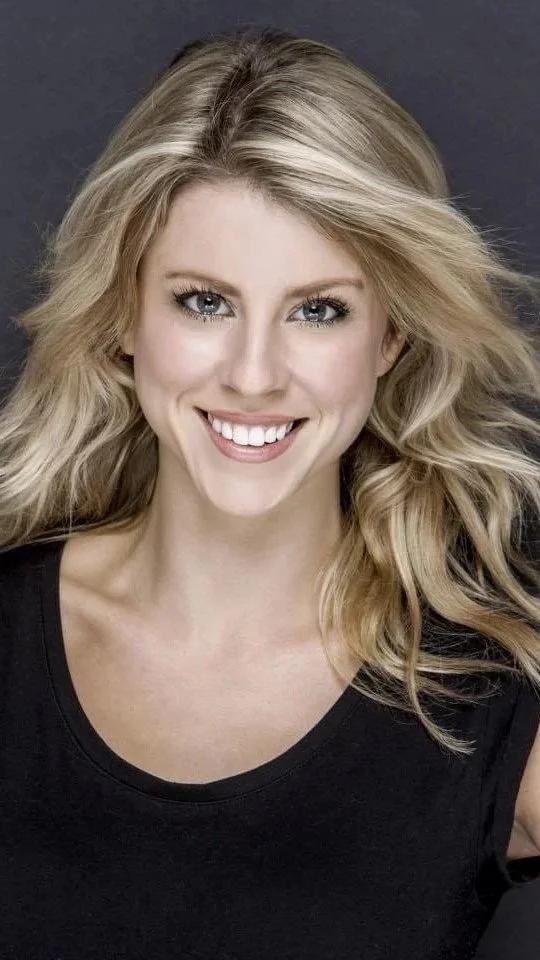 A woman with blonde hair styled in loose waves, smiling, wearing a black top, against a dark gray background.