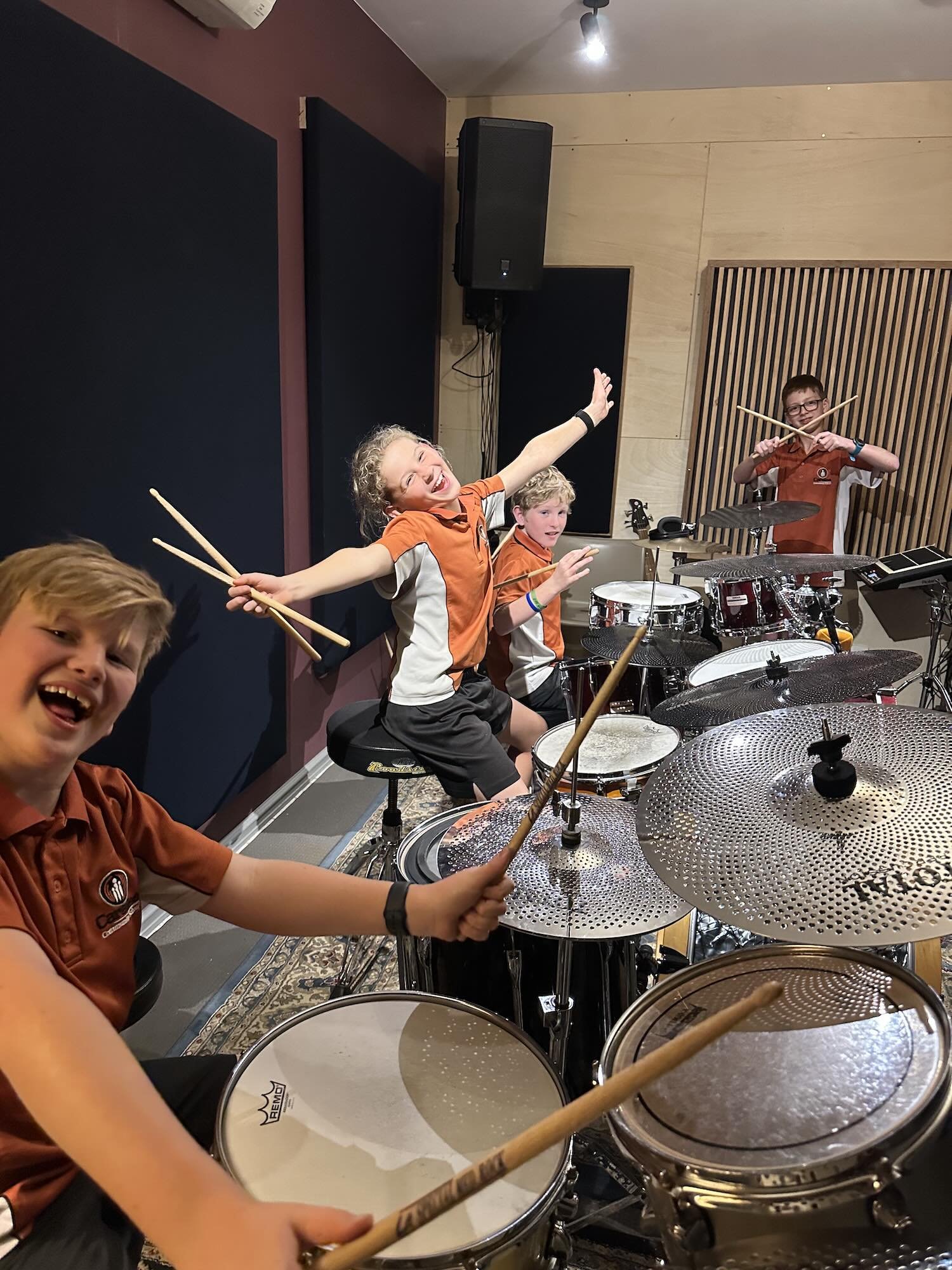 Children holding drumsticks beside drum kits during a group lesson.