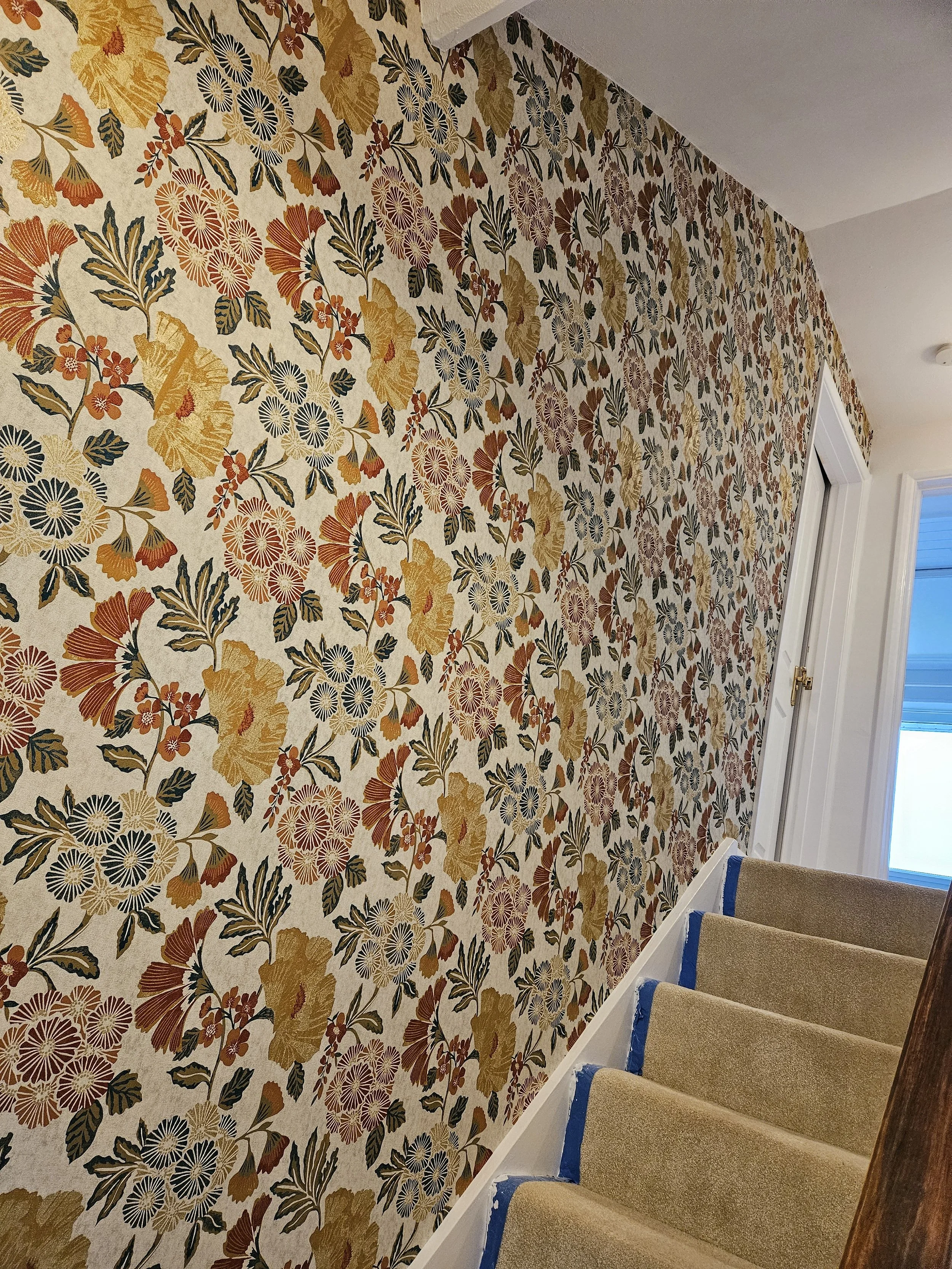 Interior staircase with beige carpeted steps and a floral decorated wall with yellow, red, purple, and black flowers and leaves.