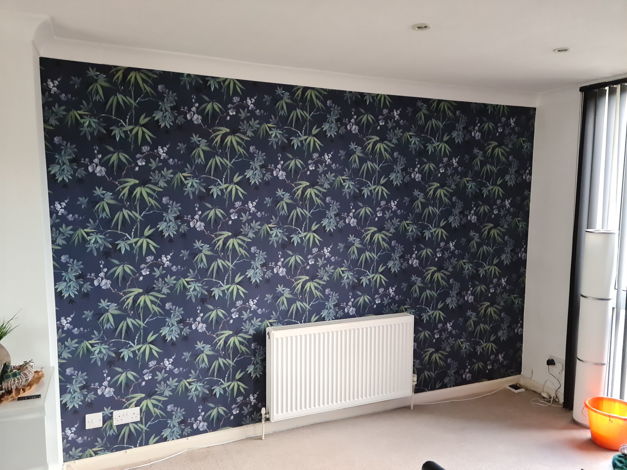 Living room with navy blue accent wall featuring tropical green leaves and white flowers, white radiator, electrical outlets, and a corner with a red-orange bowl and a tall white fan.
