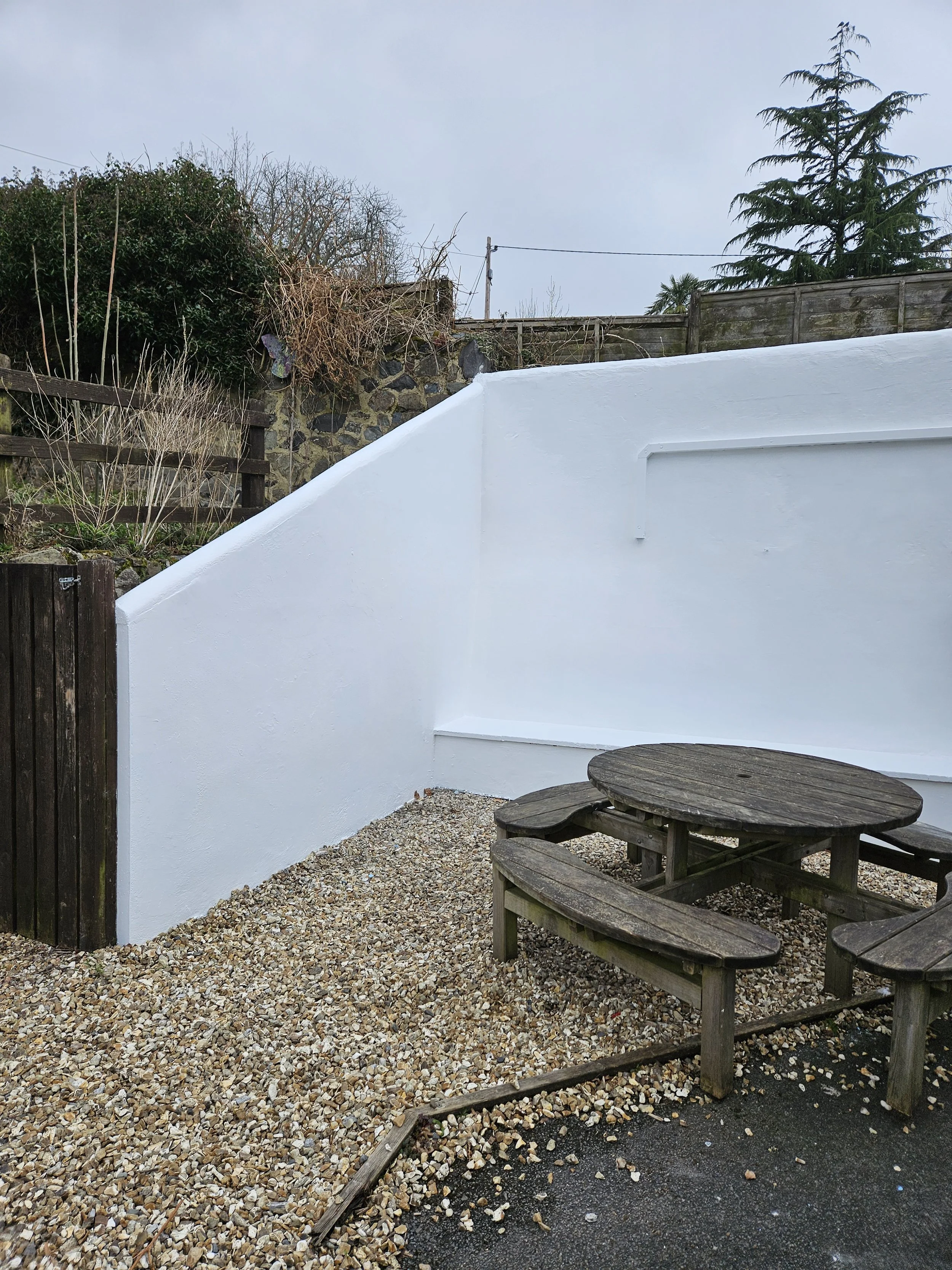 A small outdoor area with gravel ground, a wooden picnic table with attached benches, surrounded by a white painted wall on one side. There is a wooden fence and some trees and bushes in the background.