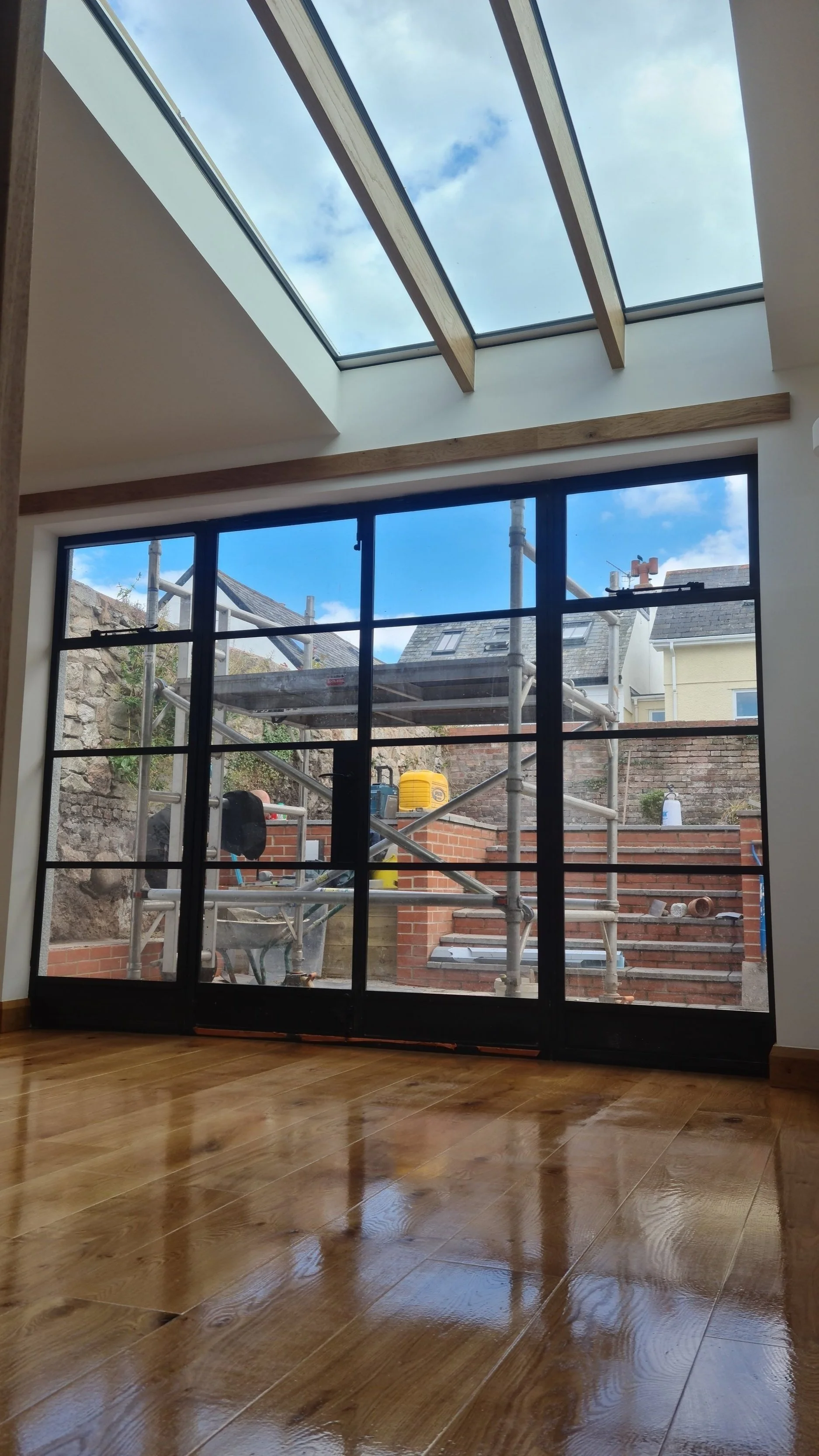 Interior view of a room with wooden flooring, large glass sliding doors, and a skylight ceiling, showing an outdoor construction scene with scaffolding and tools.