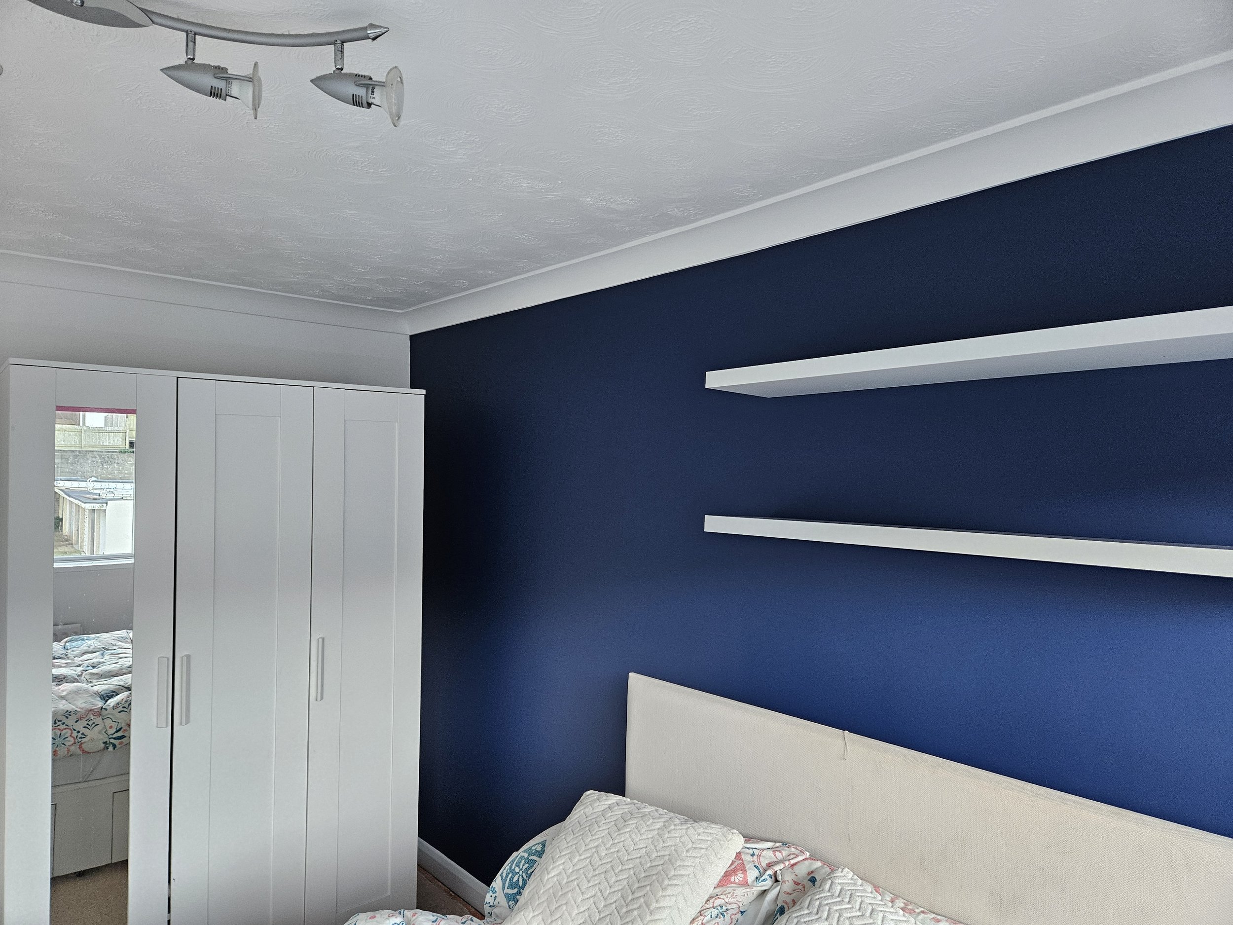 A bedroom with a navy blue accent wall, white shelves, a white wardrobe with a mirror, and a bed with white and patterned bedding.