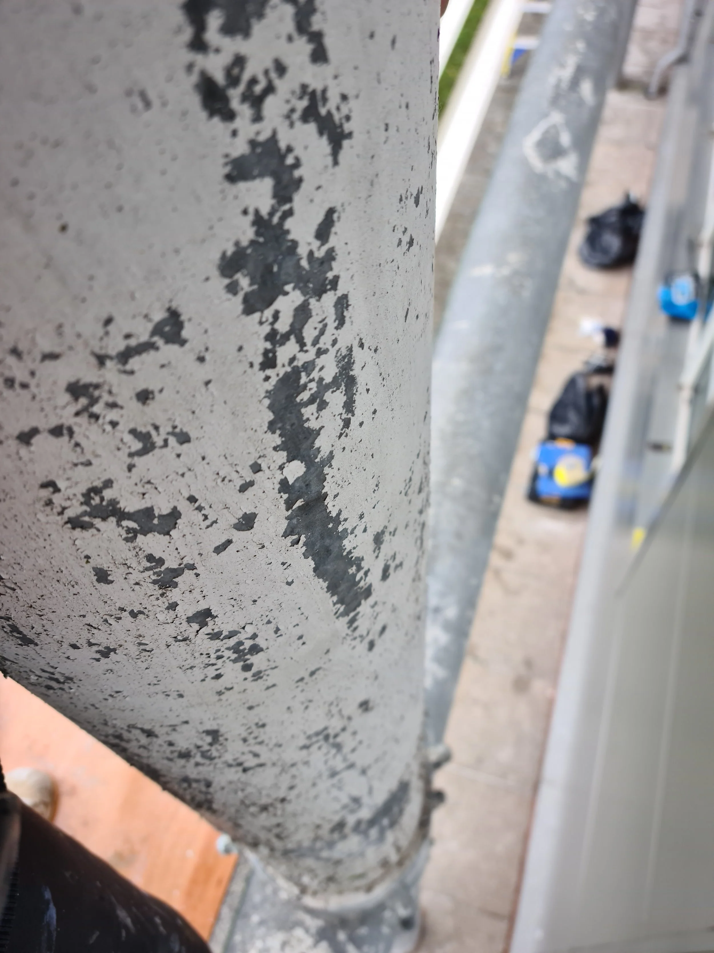 Close-up of a weathered metallic pole with peeling gray and black paint, with a blurred background showing some bags, a broom, and a leash on the ground.