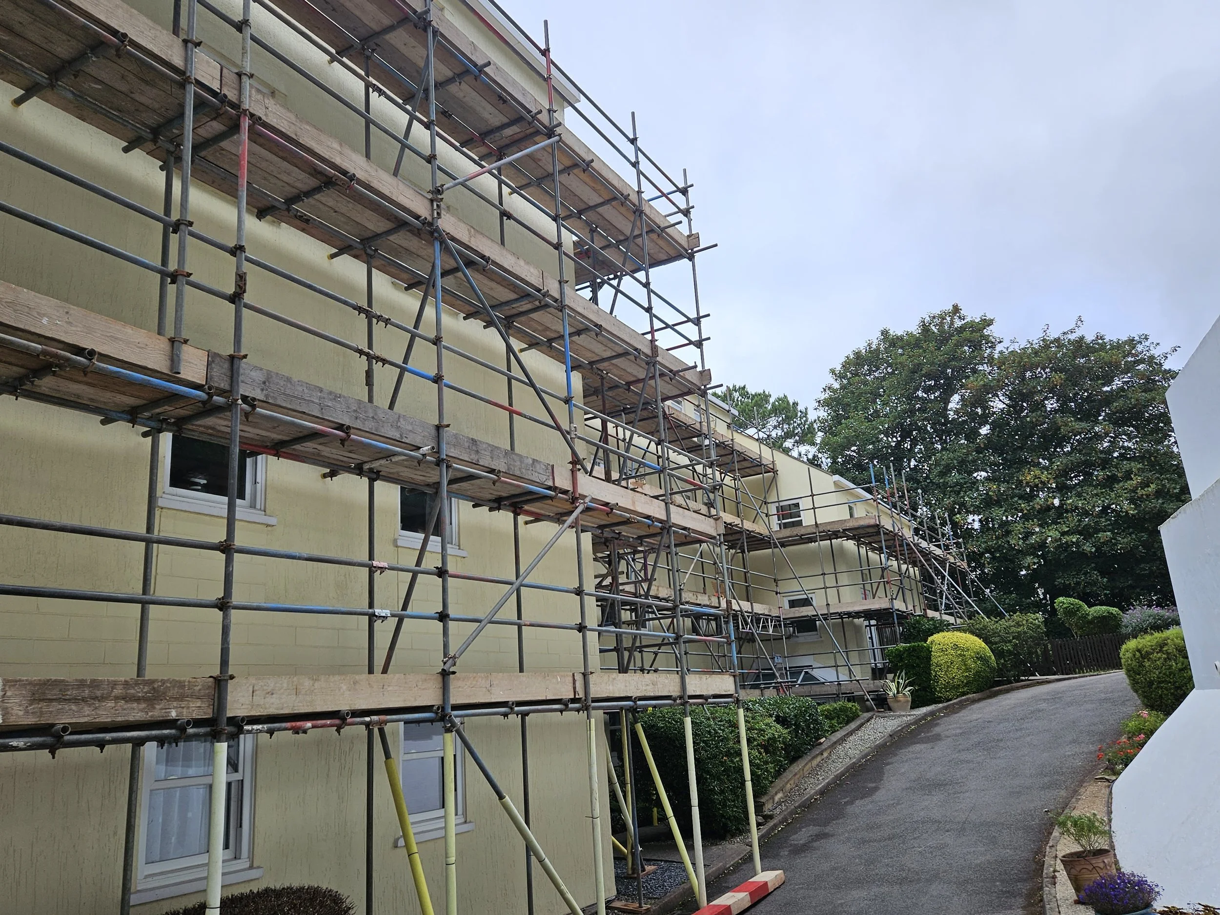 Construction scaffolding surrounding a yellow building along a curved driveway, with trees and shrubs in the background.