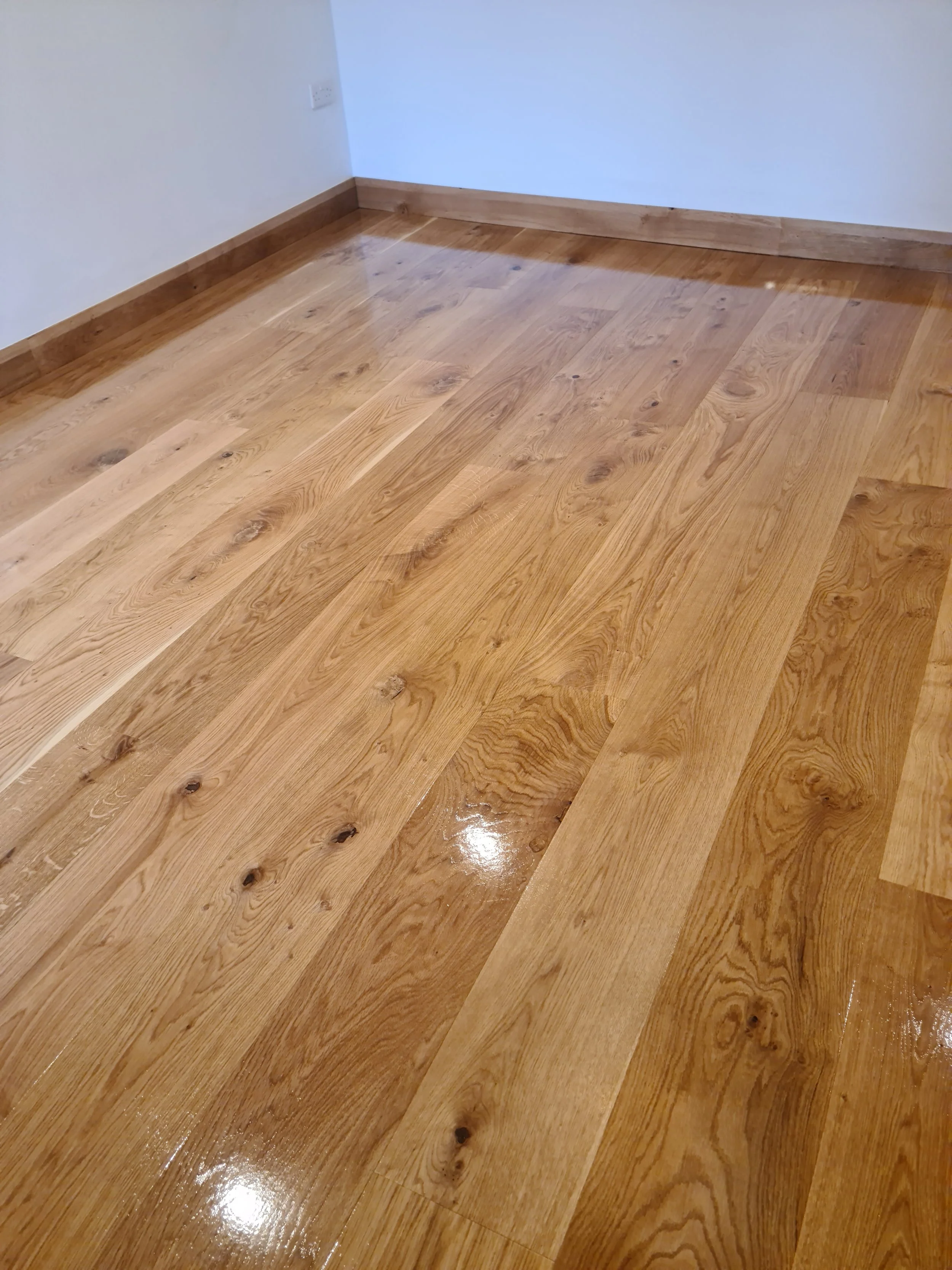 Polished hardwood floor with visible wood grain and knots in a corner of a room with white walls and wood baseboards.
