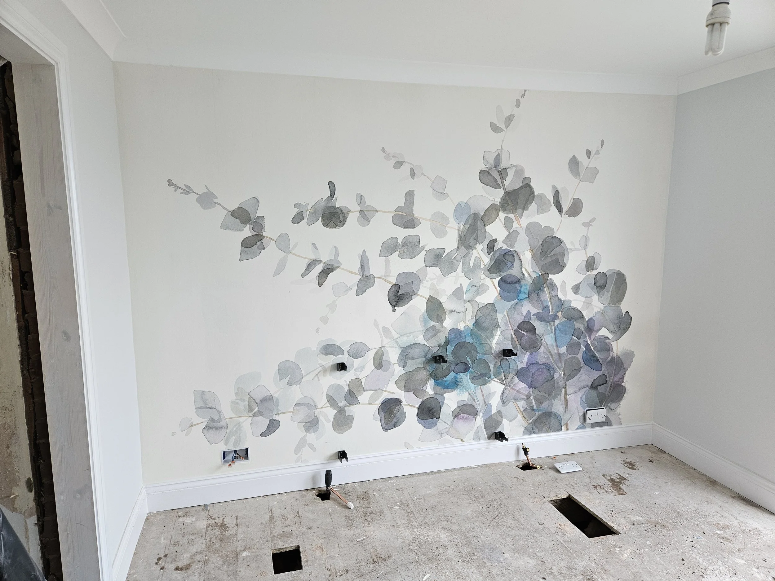Partially finished room with a white wall decorated with a watercolor style mountain ash leaf pattern mural, electrical outlets, and holes in the floor for plumbing or wiring.