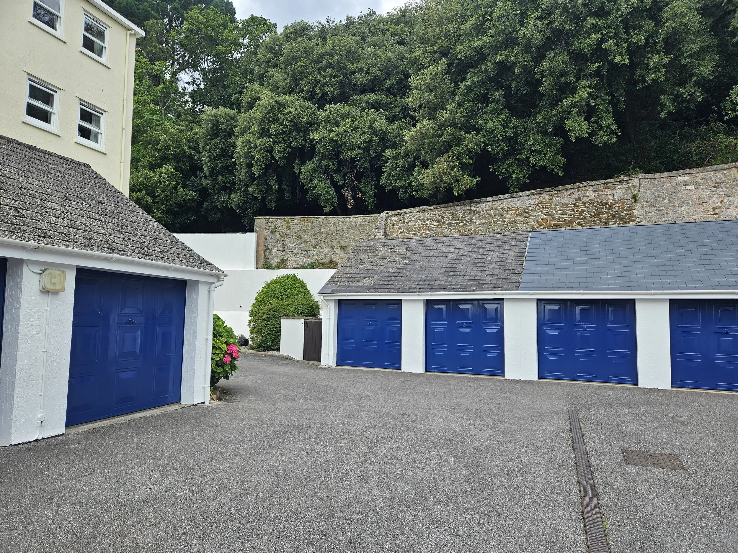 Multiple closed garages with blue doors, surrounded by white walls and a paved driveway, with trees and a stone wall in the background.