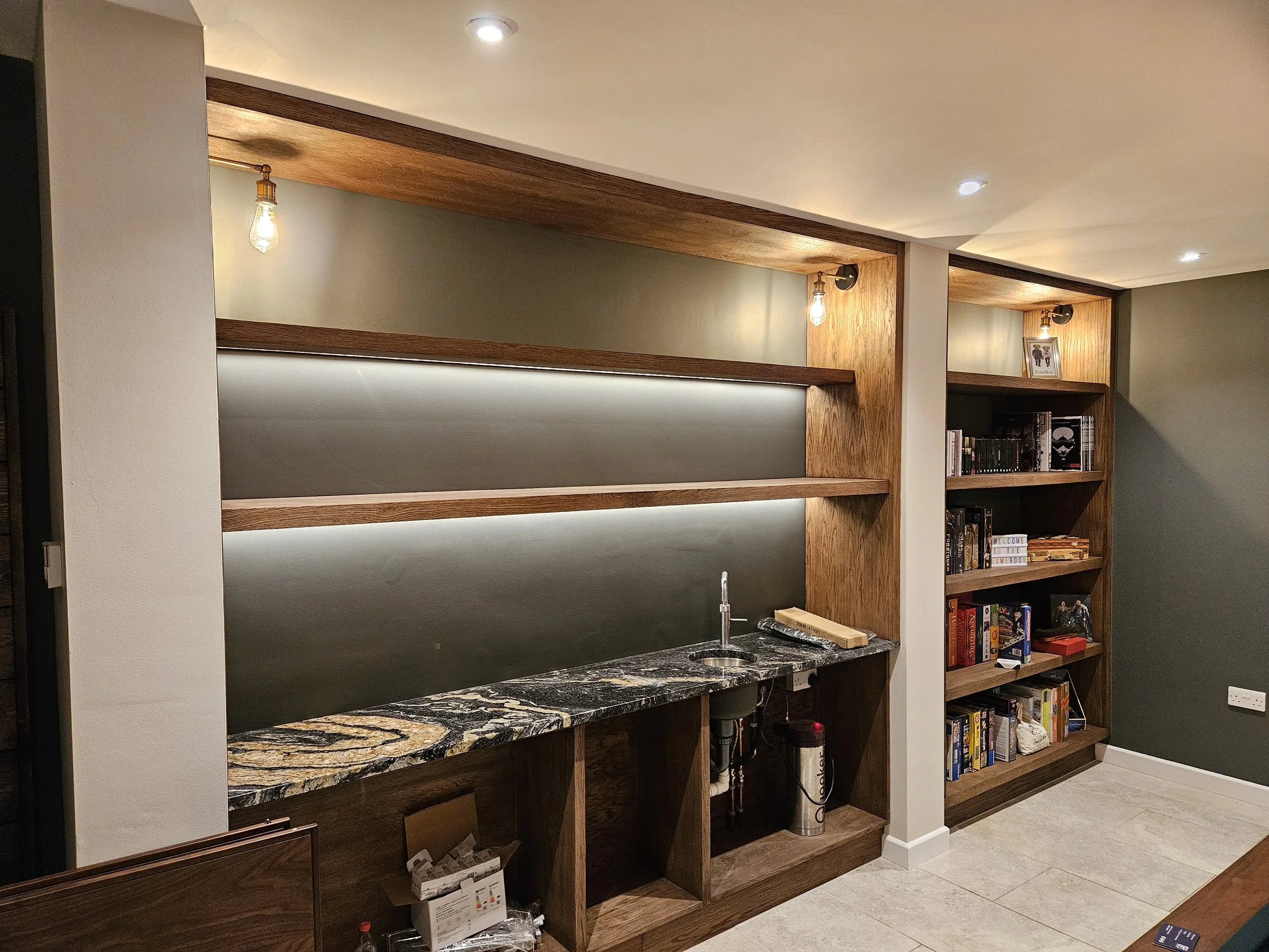 Living room with built-in wooden shelves, a marble countertop with a sink, a small bookshelf filled with books, and warm lighting fixtures.