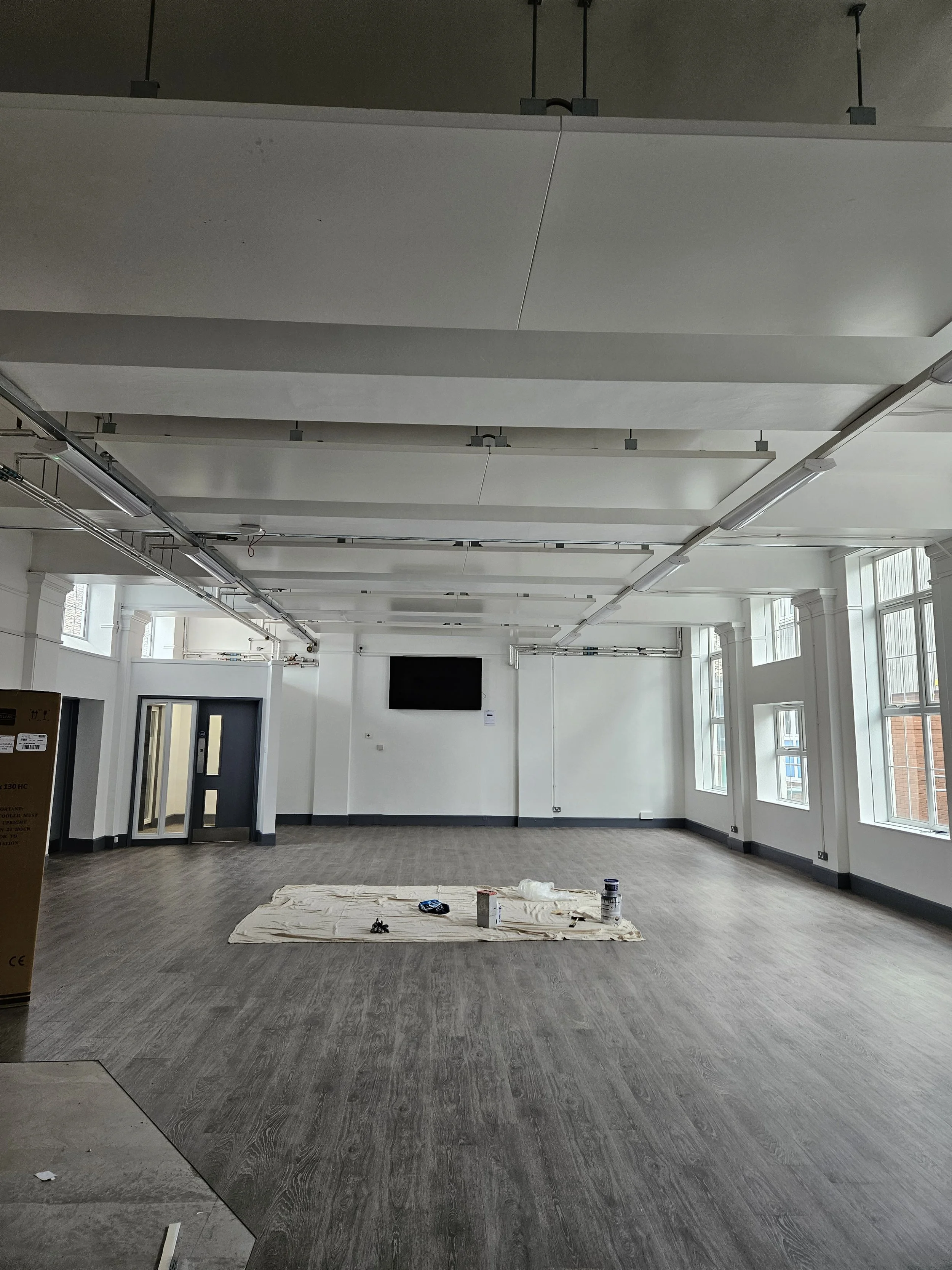 Empty room with large windows, a mounted TV on the wall, and painting supplies on a sheet on the floor.