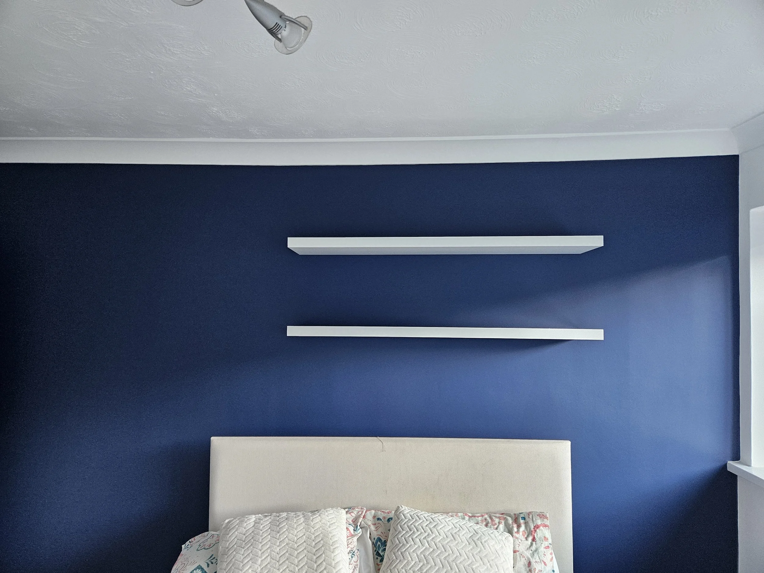 A bedroom with a navy blue wall decorated with two white floating shelves. Part of a bed with a white headboard and pillows is visible at the bottom of the image. The ceiling has a light fixture.