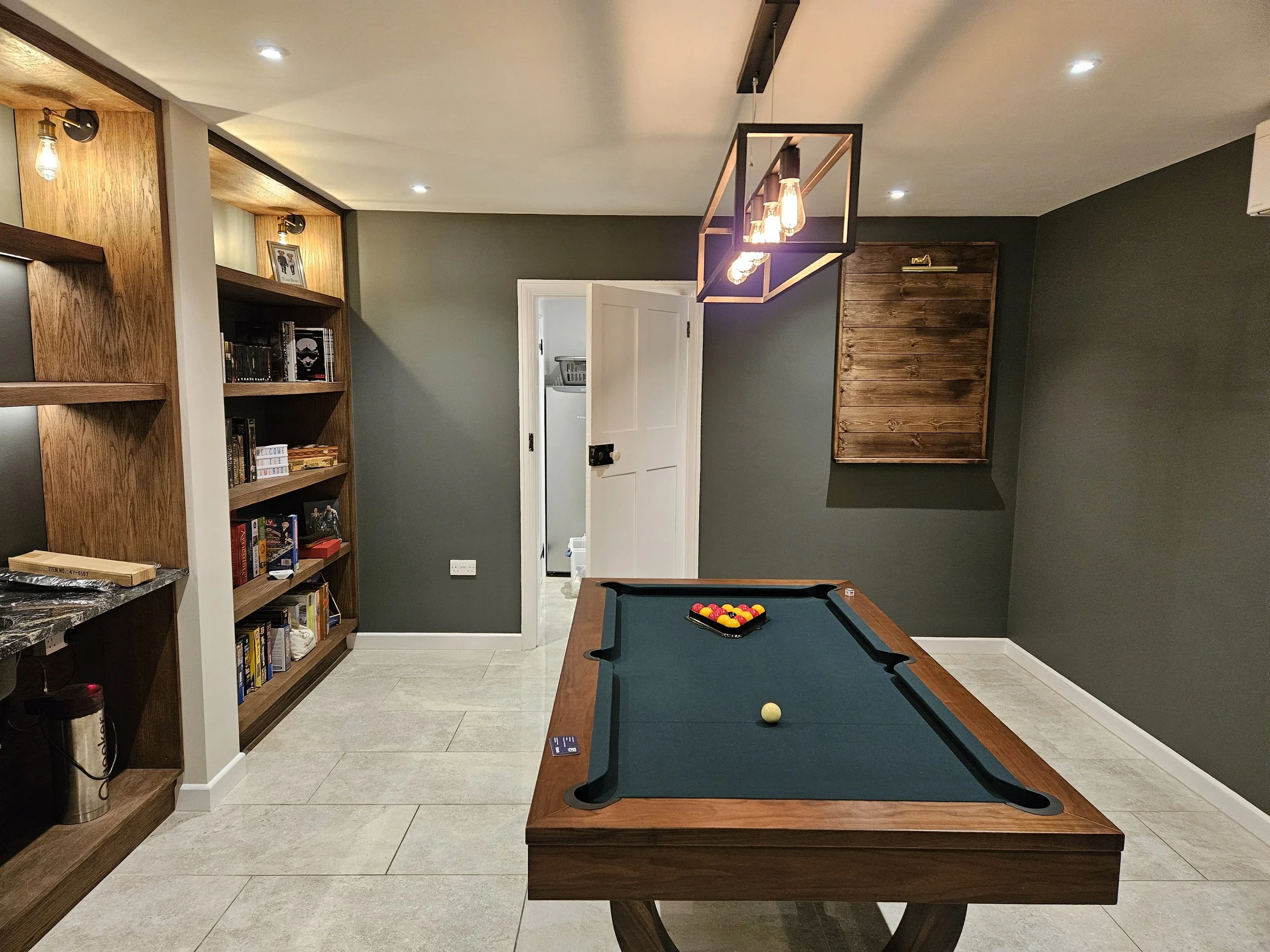 A home game room with a pool table in the center, a bookshelf filled with books on the left, and a wooden wall panel on the right. A modern light fixture hangs above the pool table, and there are small ceiling lights in the room.