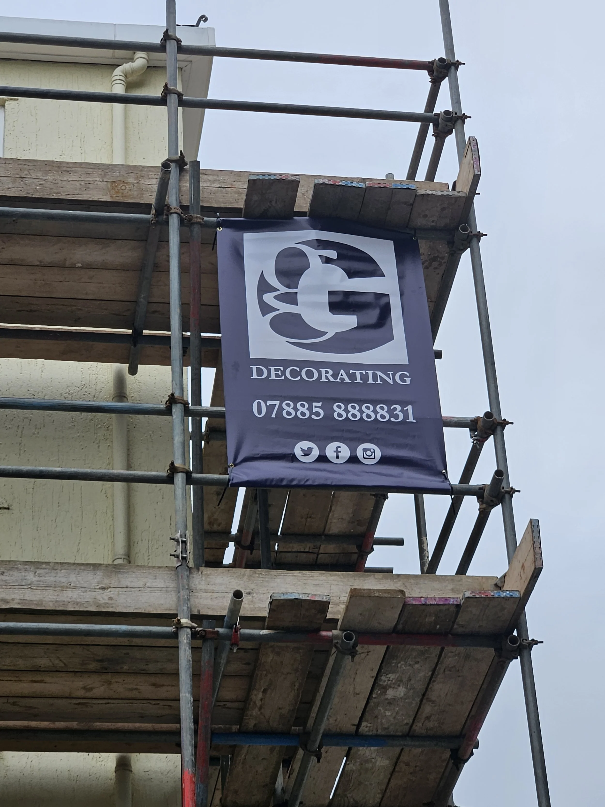 Scaffolding with a banner displaying a logo, contact phone number, and social media icons for a decorating business.