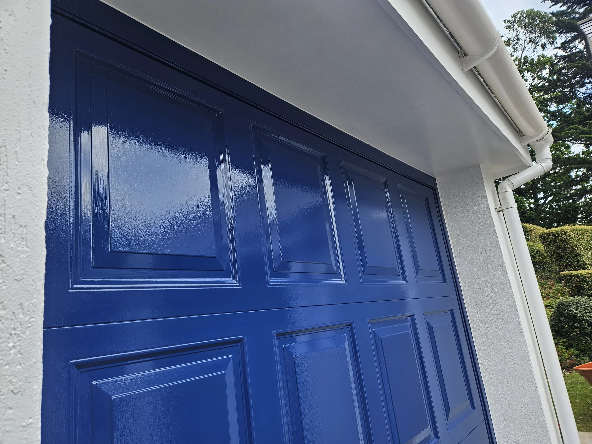 Close-up of a blue garage door with a textured white wall on the left and white rain gutter pipes on the top right, with green trees and hedges in the background.