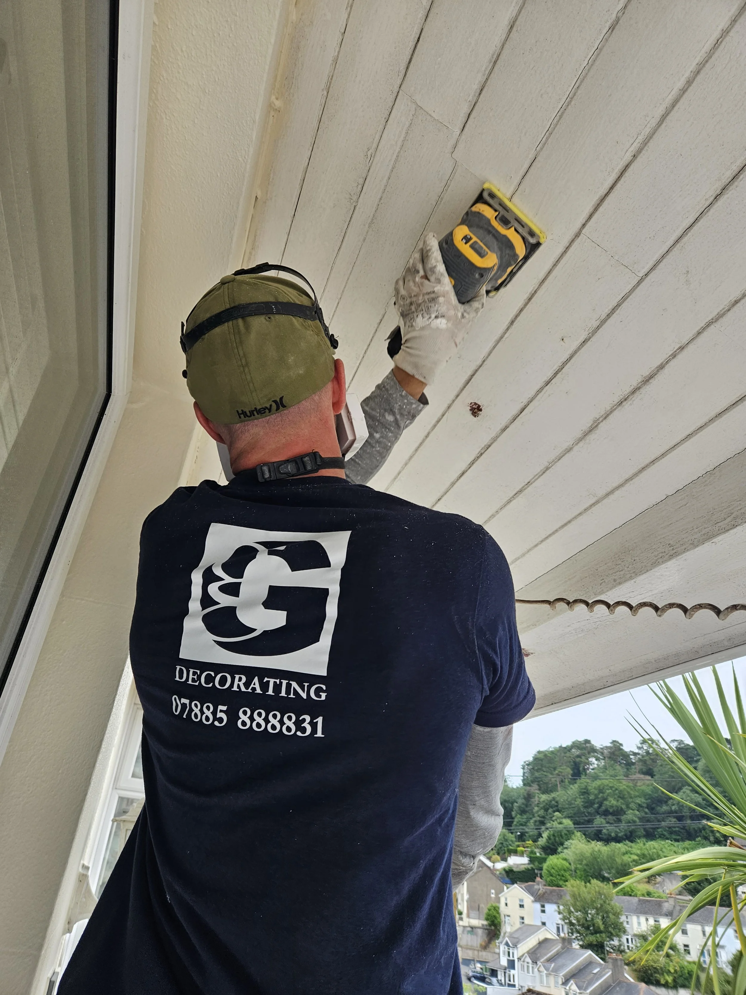 A man wearing a black t-shirt with the words 'Decorating' and a phone number on the back, an olive green cap, and white gloves is sanding a white wooden outdoor ceiling with a yellow sander.