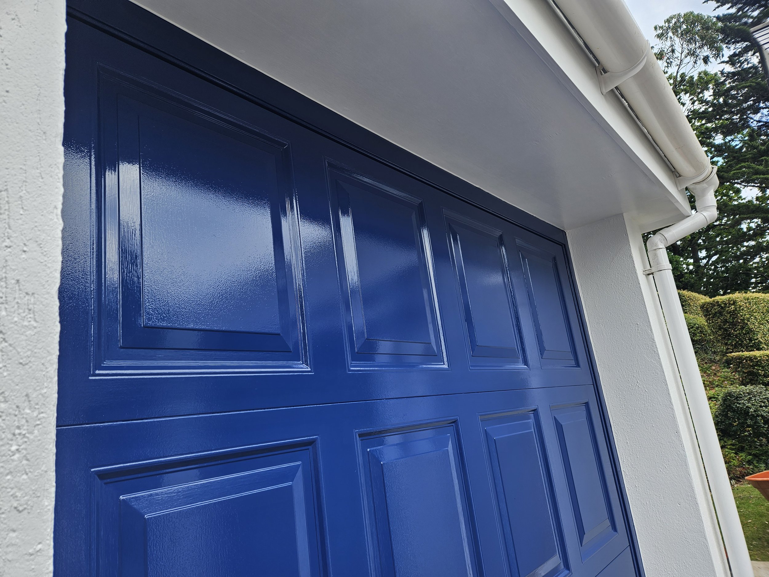 Close-up of a bright blue garage door with decorative panels, white textured walls, and white gutter pipes, under a gray fascia board.