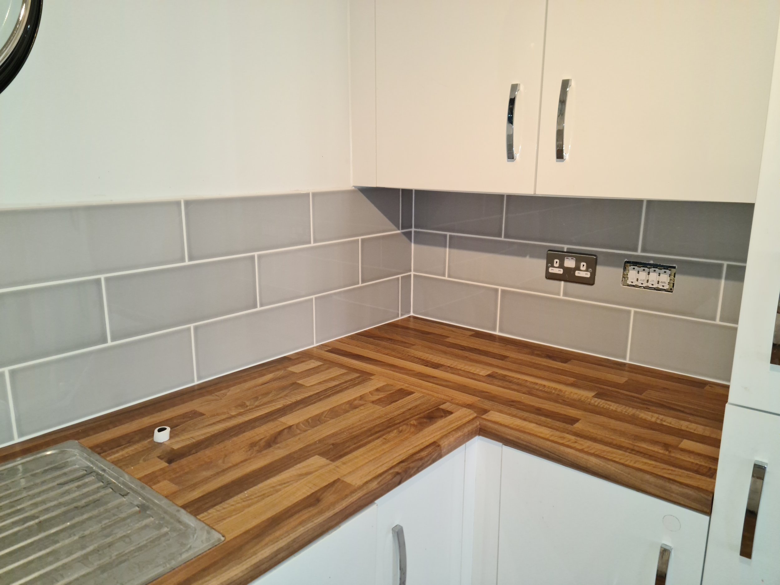 Kitchen countertop with wooden surface, gray tiled backsplash, white cabinets, with an electrical outlet and a socket near the corner.