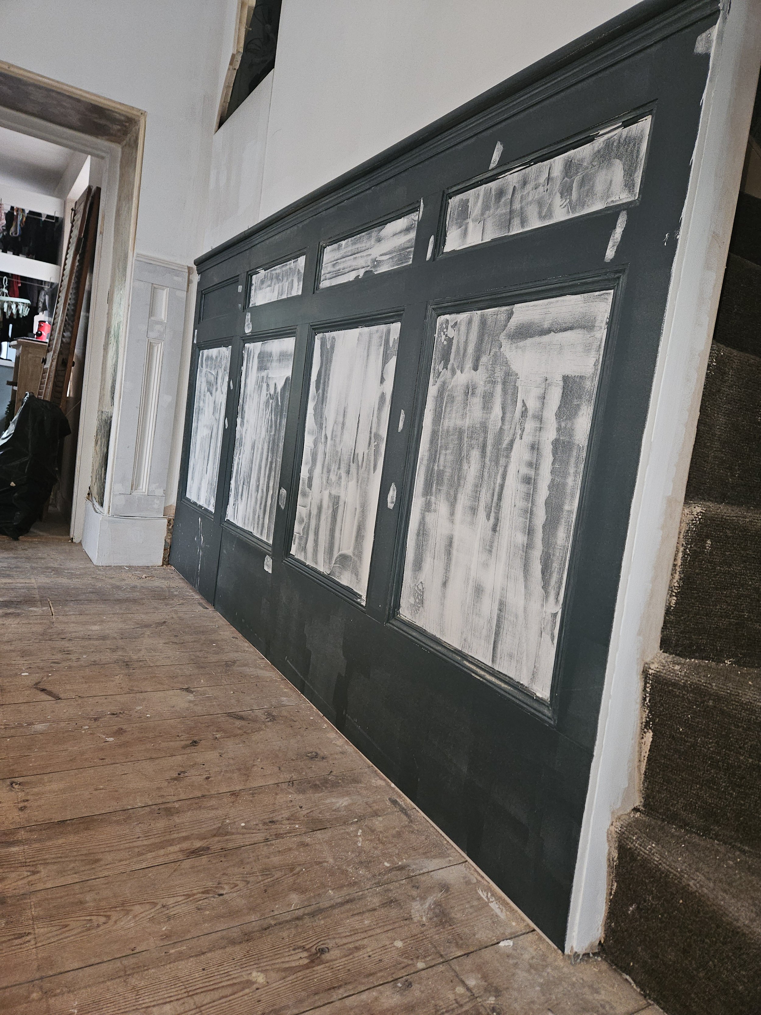Interior of a room with a large black-paneled wall or furniture piece under construction, with visible areas of white primer or paint, next to a staircase with brown carpeted steps and a hardwood floor that has some dust and debris.