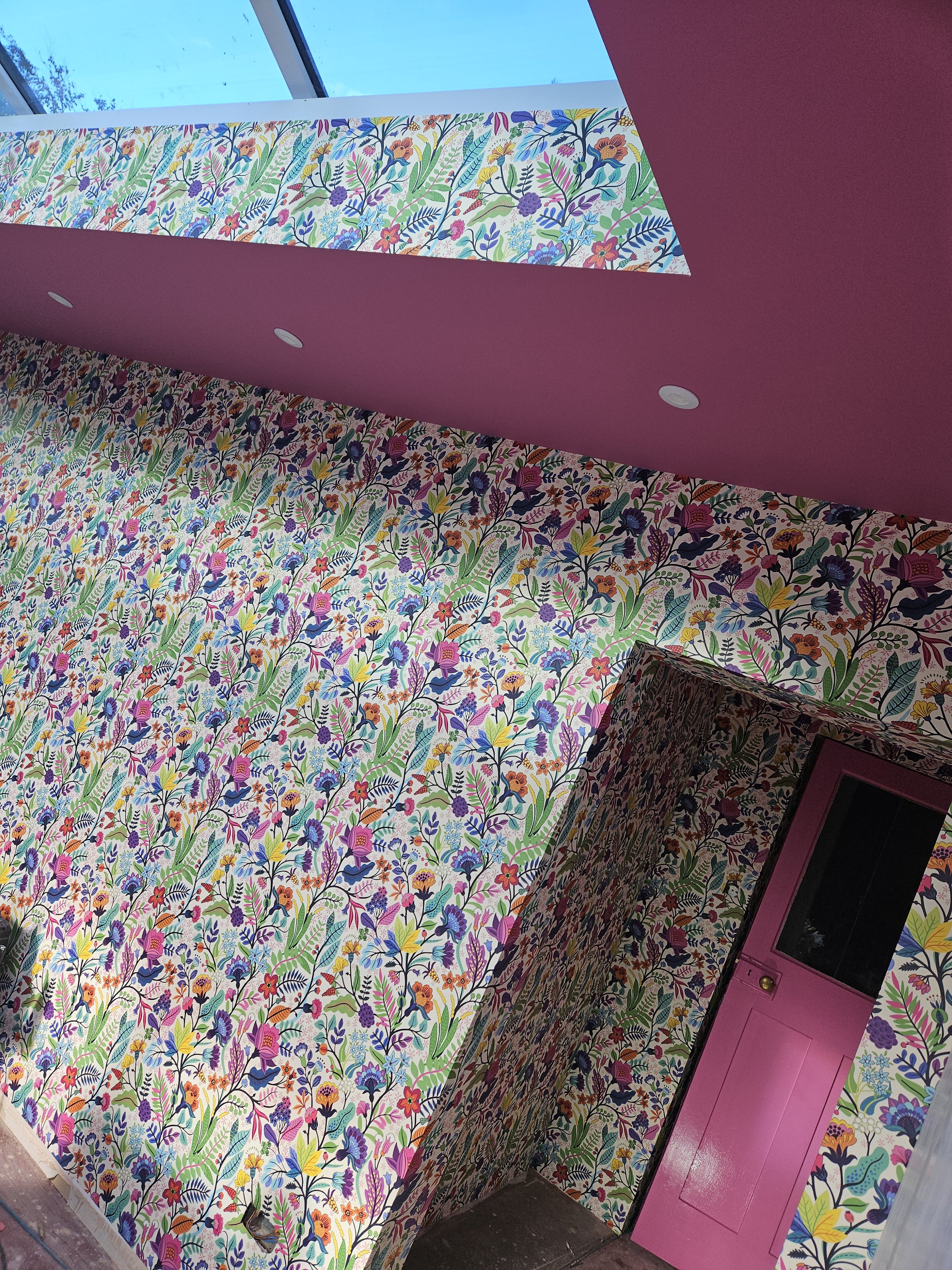 A room with walls and a door decorated in colorful floral wallpaper and a pink ceiling with a skylight.