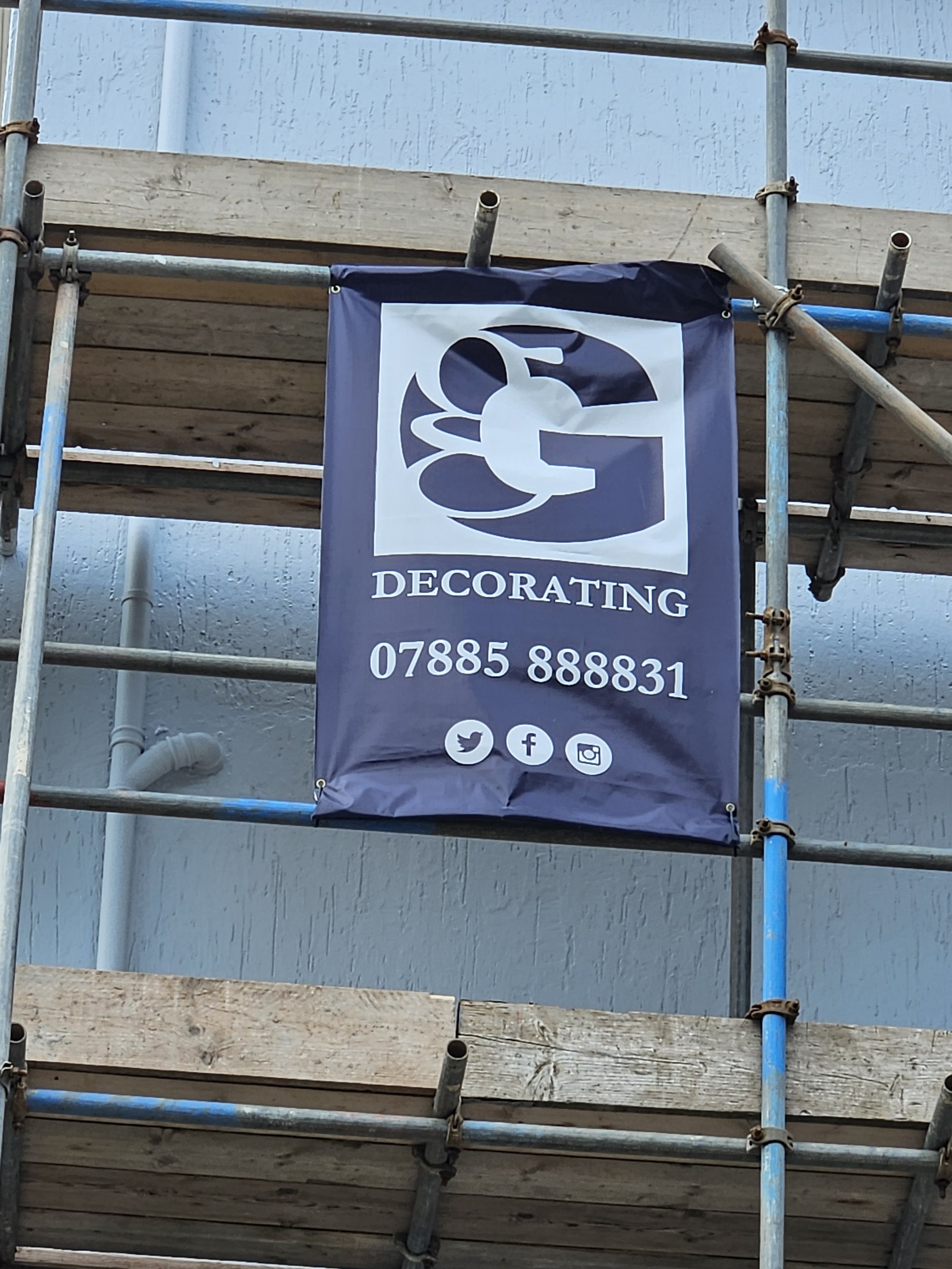 Banner for a decorating business displayed on scaffolding, with contact number and social media icons.