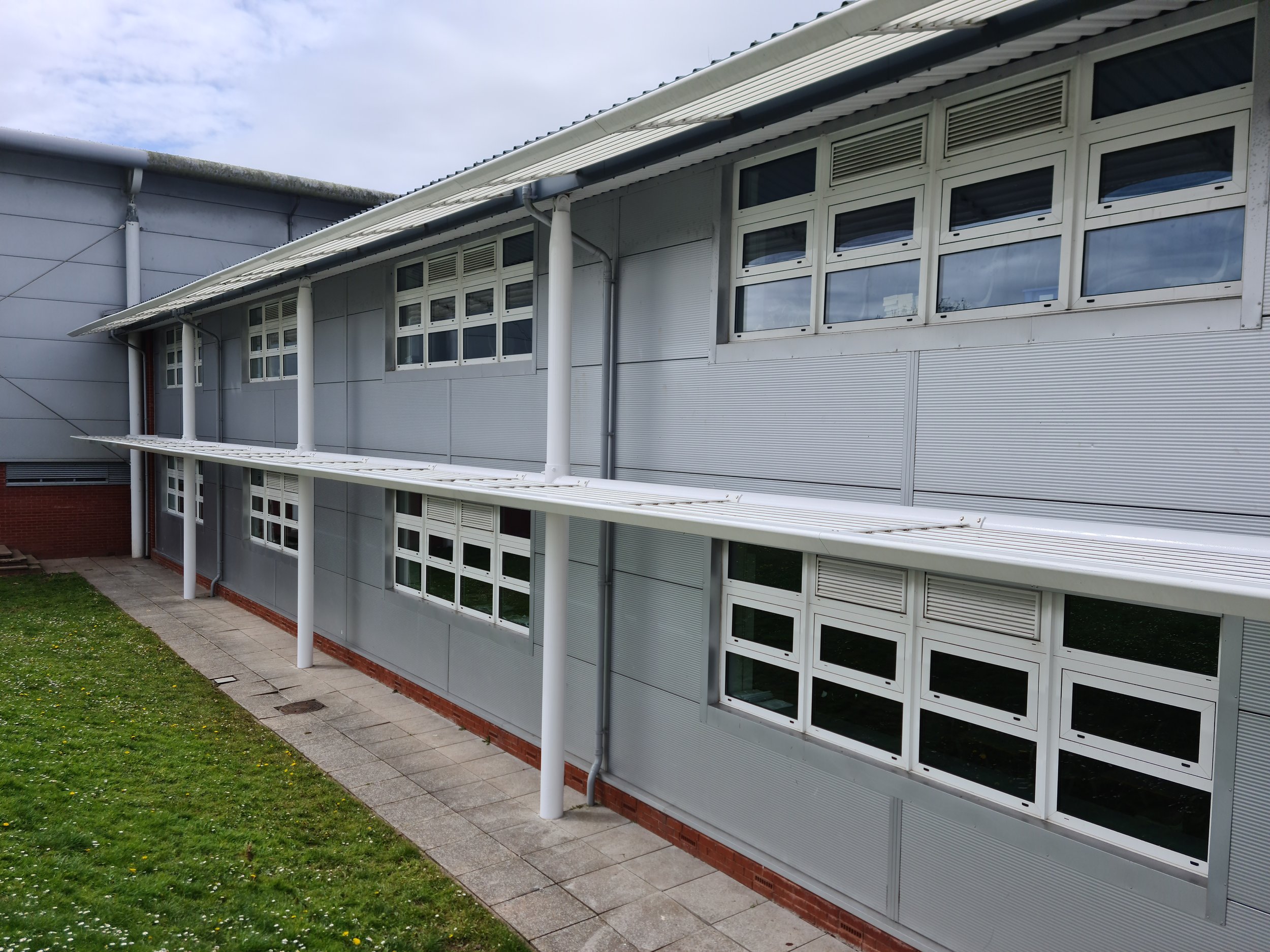 Exterior of a modern school building with a gray facade, multiple PVC windows, and a small grassy area with a pathway.