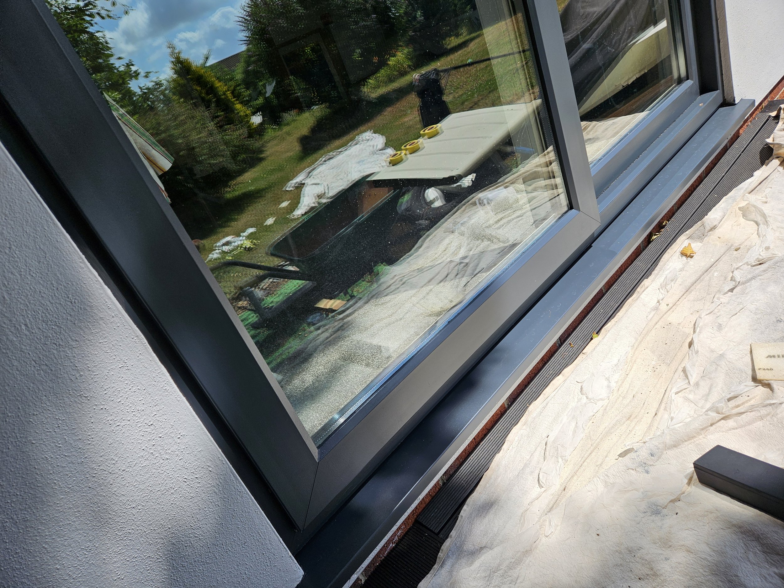 View of a newly installed large window with a black frame on a building exterior, with construction tools and paper covering the ground outside.