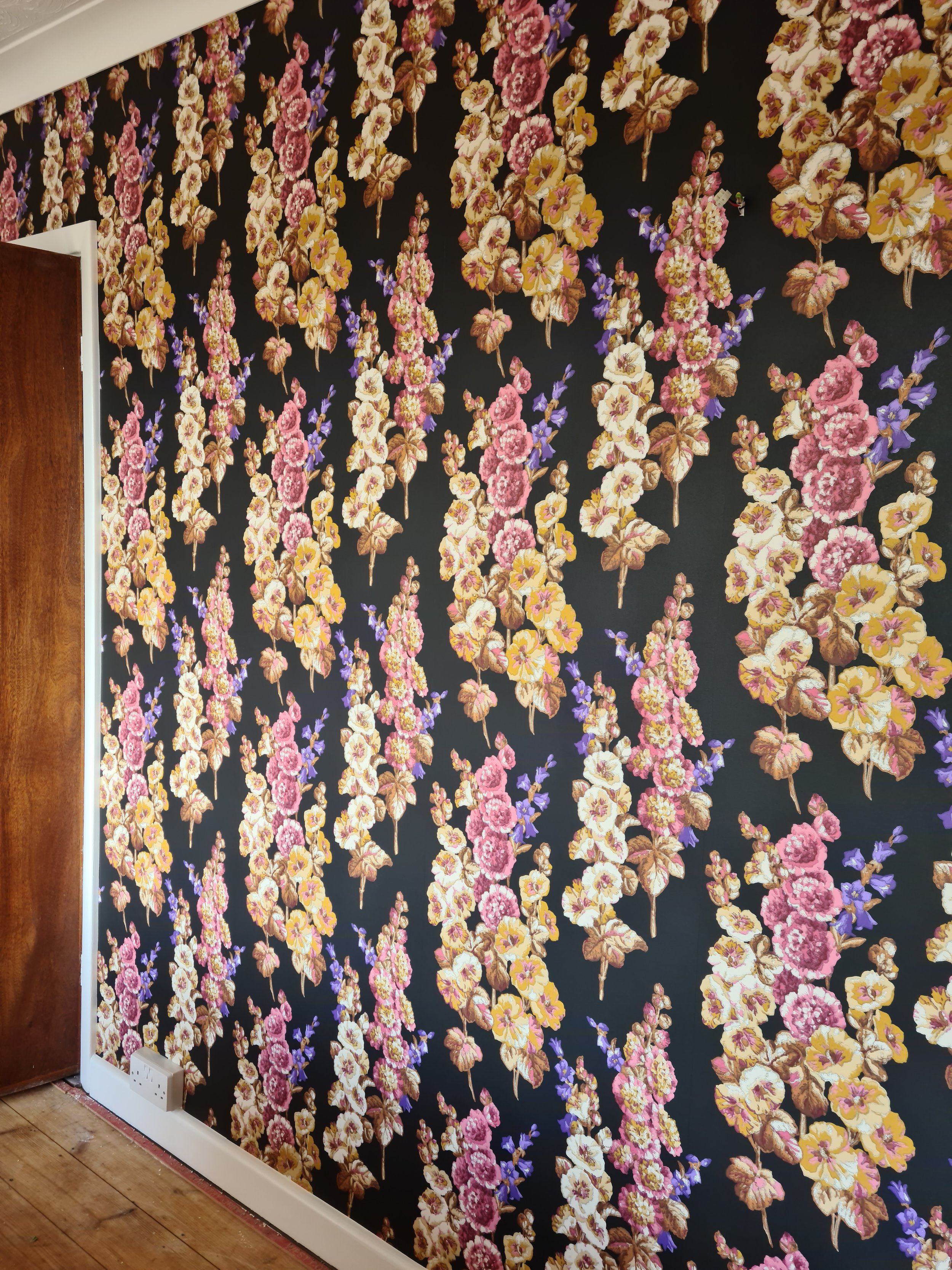 A wall covered with dark wallpaper featuring a pattern of hanging floral bouquets in pink, yellow, white, and purple, with electrical outlets at the bottom and a wooden door on the left side.