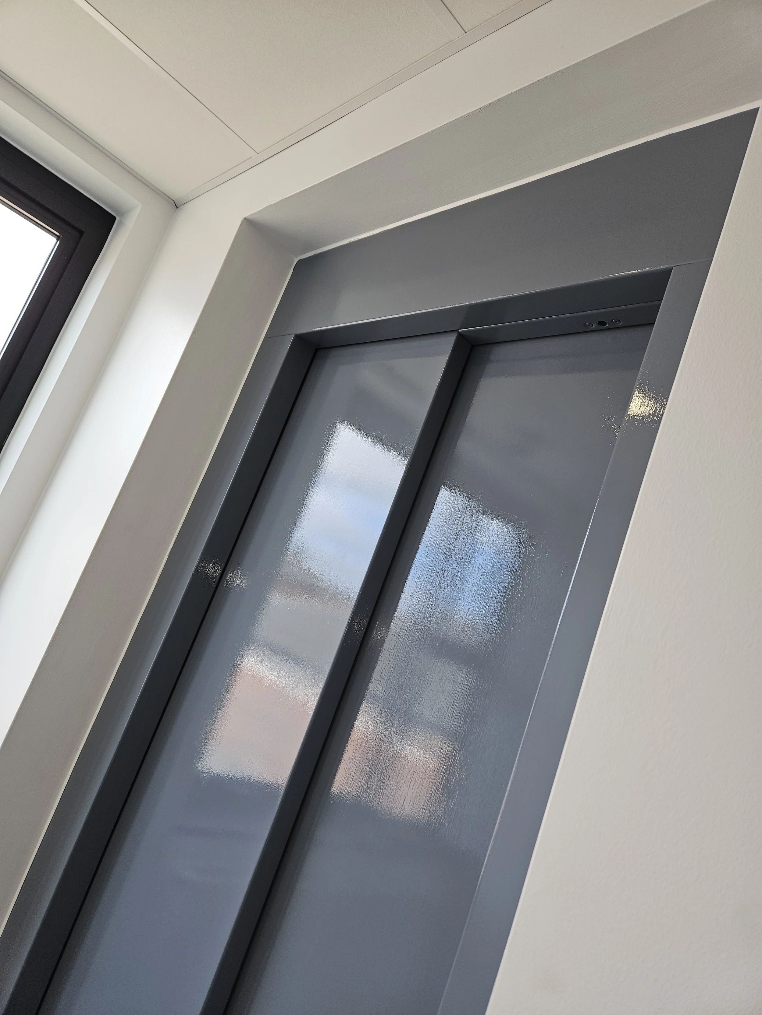 A closed elevator door painted in gray color in a modern building interior with white walls and ceiling, and a window nearby.